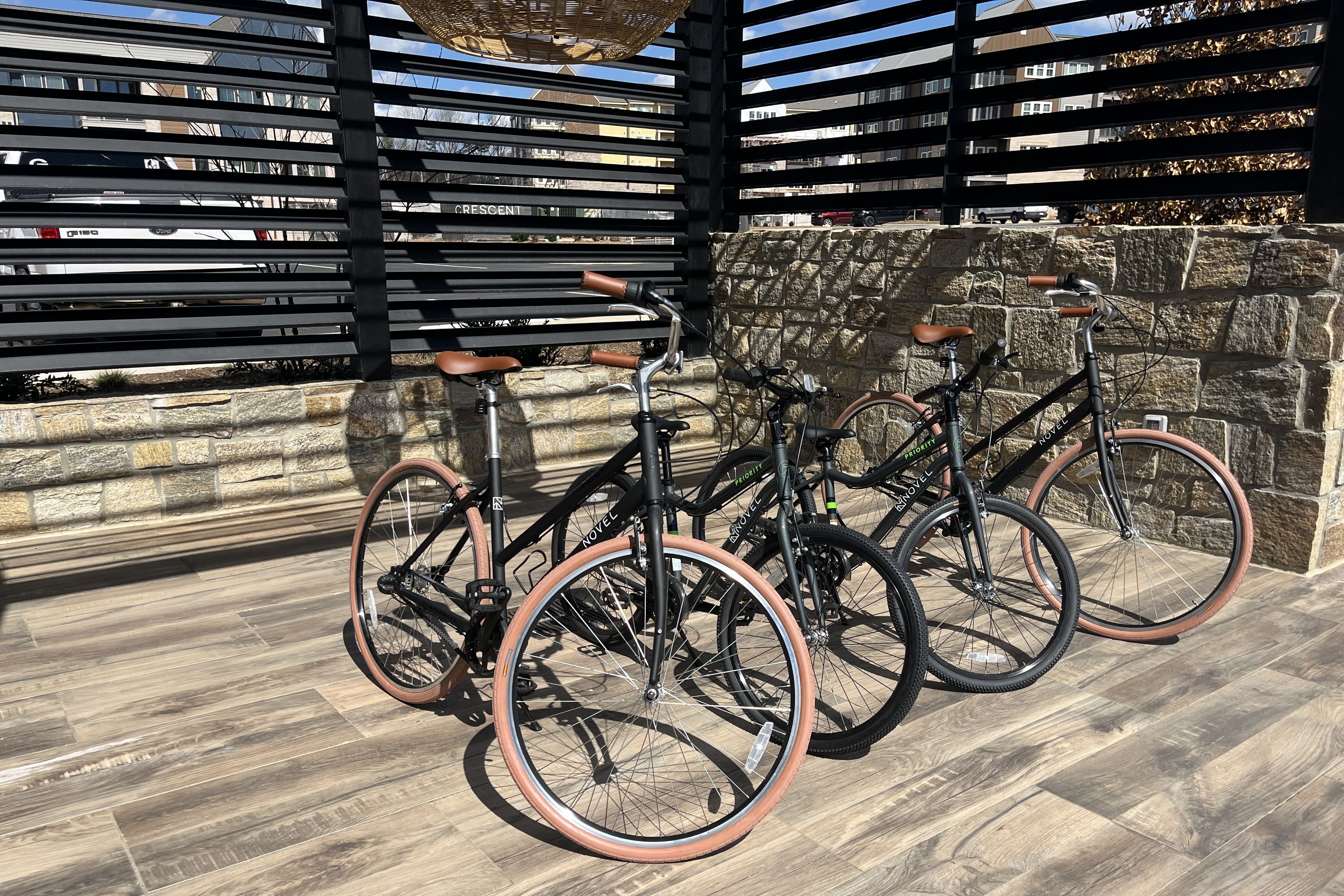 Four black bicycles with brown saddles and tan tires are parked in a row along a stone wall under a black slatted fence on a wooden-tiled patio.