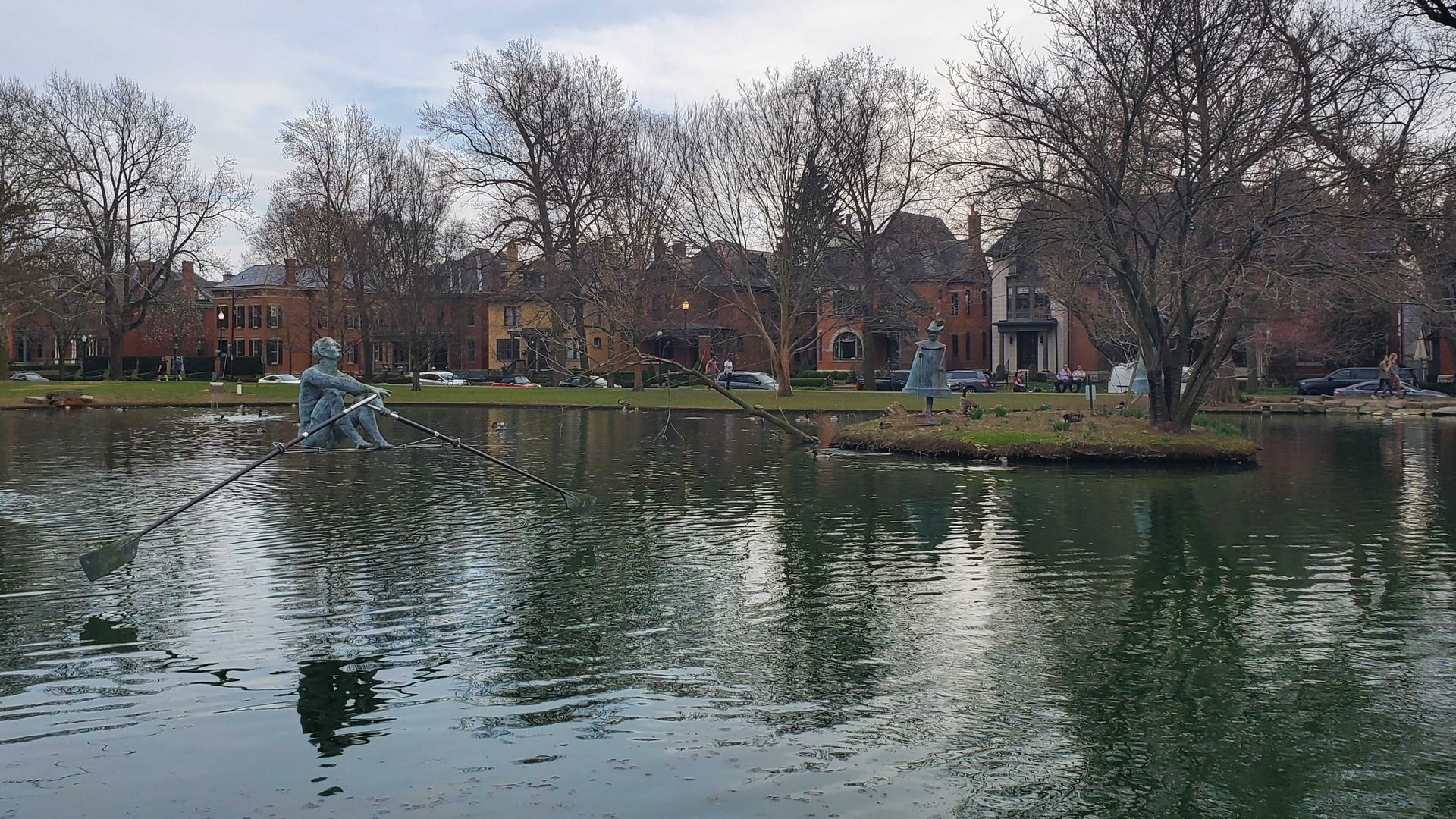 A sculpture of a rower hanging on a cable above a park lake.