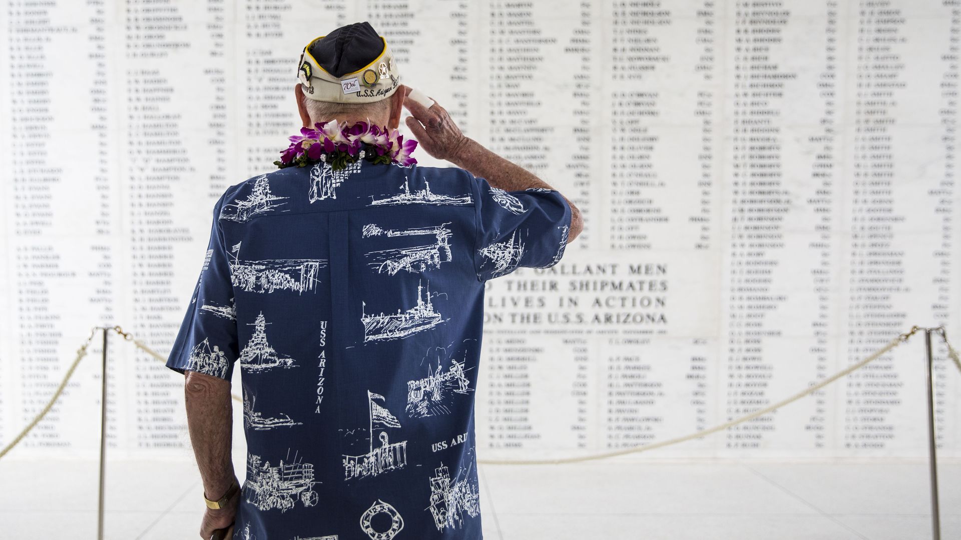 Photo shows an older man saluting a memorial wall with the names of people who died aboard the USS Arizona during the attack on Pearl Harbor