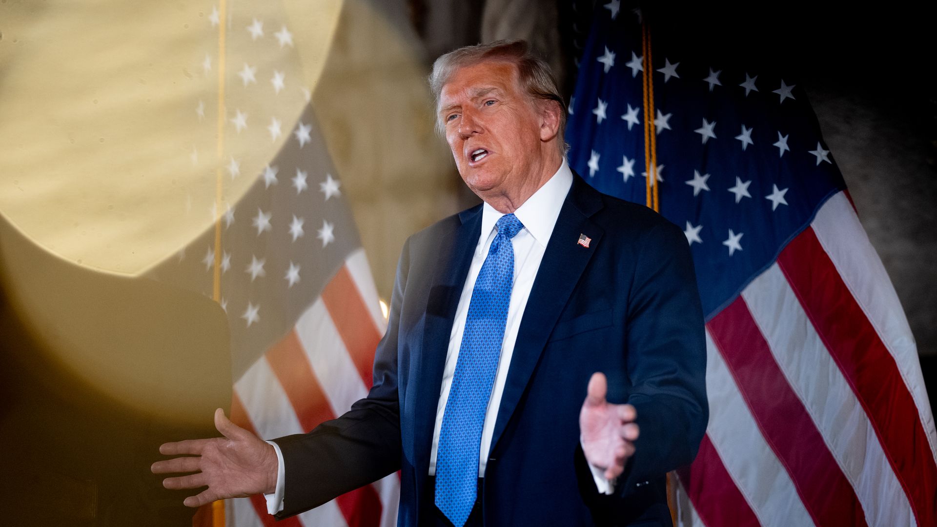 President Trump wearing a blue suit and standing in front of an American flag.