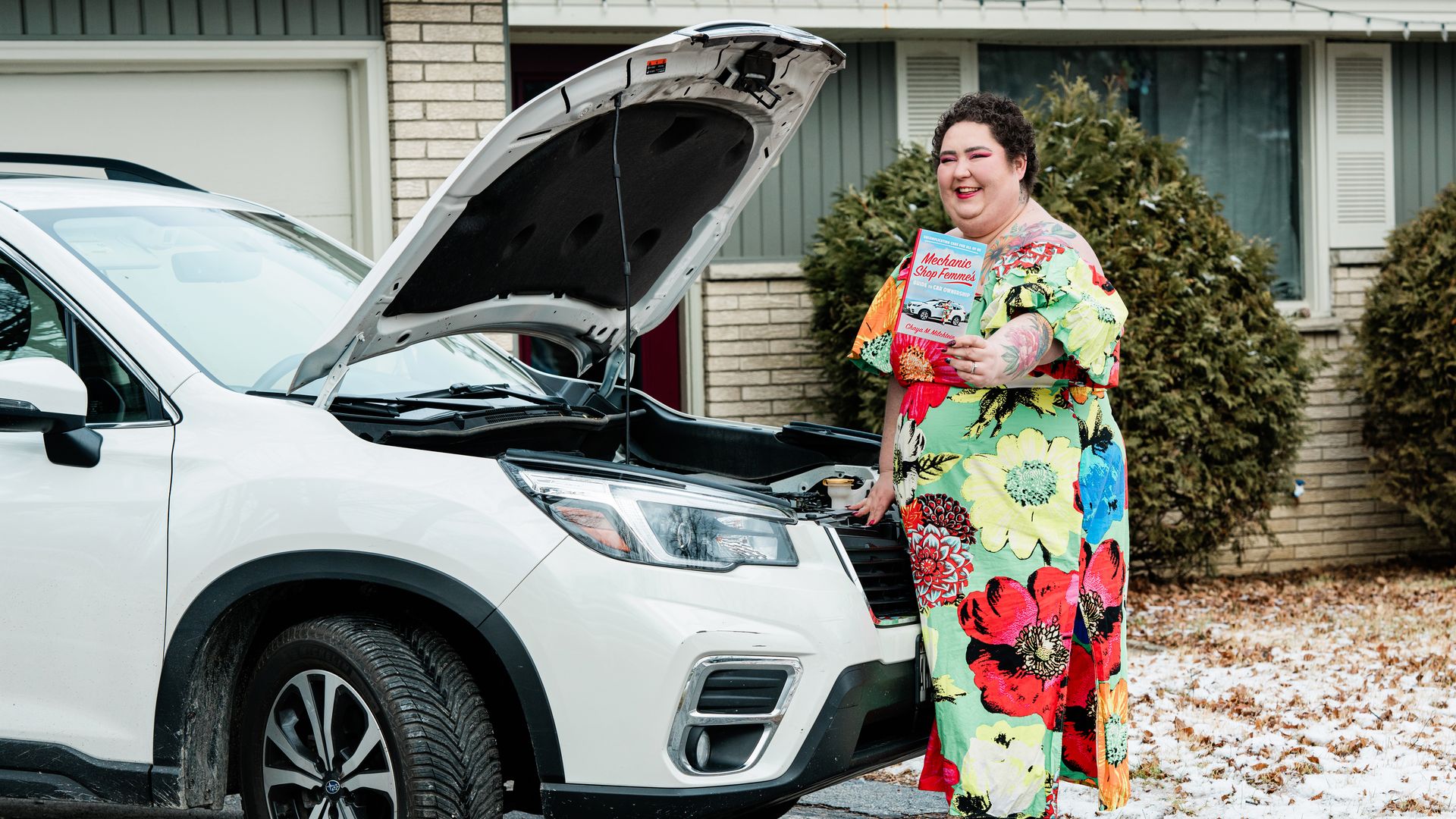 Photo of a person holding a book next to a car with its hood popped up