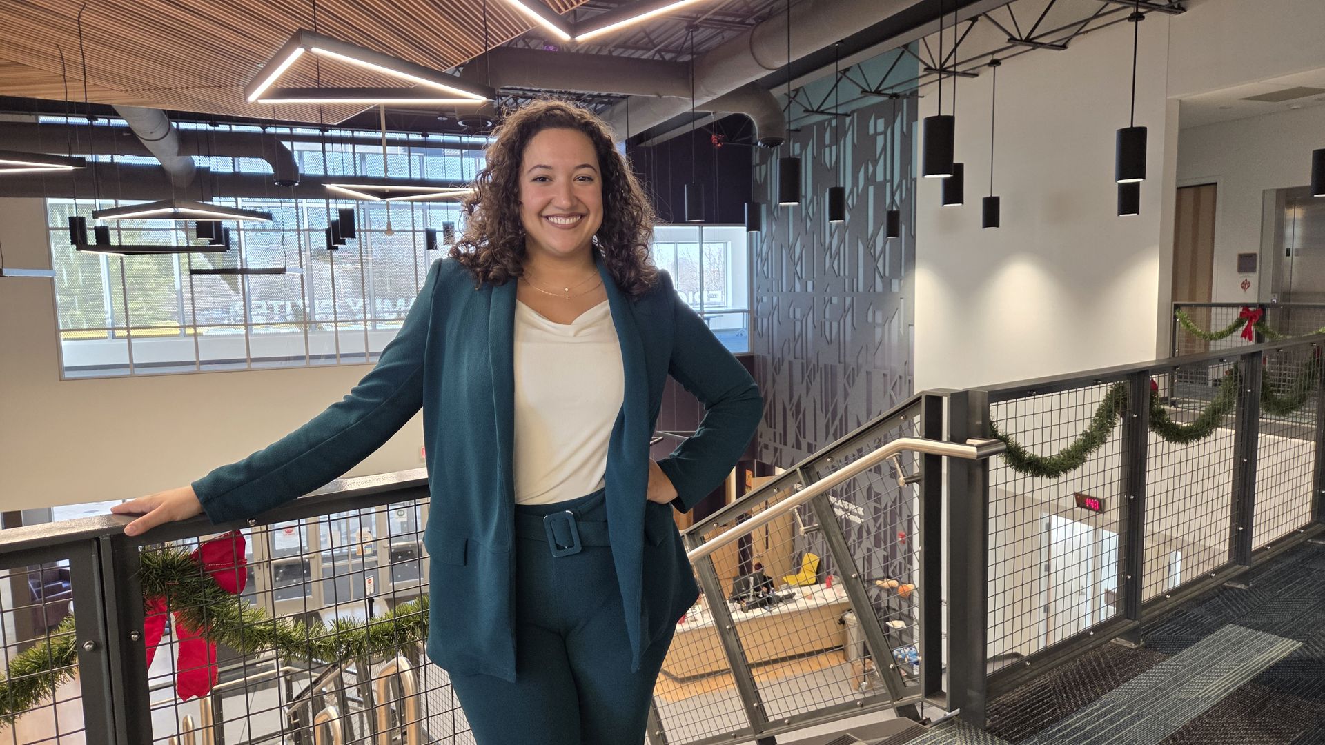 Smiling woman with curly hair wearing a dark teal suit and white top stands on a decorated indoor balcony with garlands and pendant lights, industrial ceiling in background.