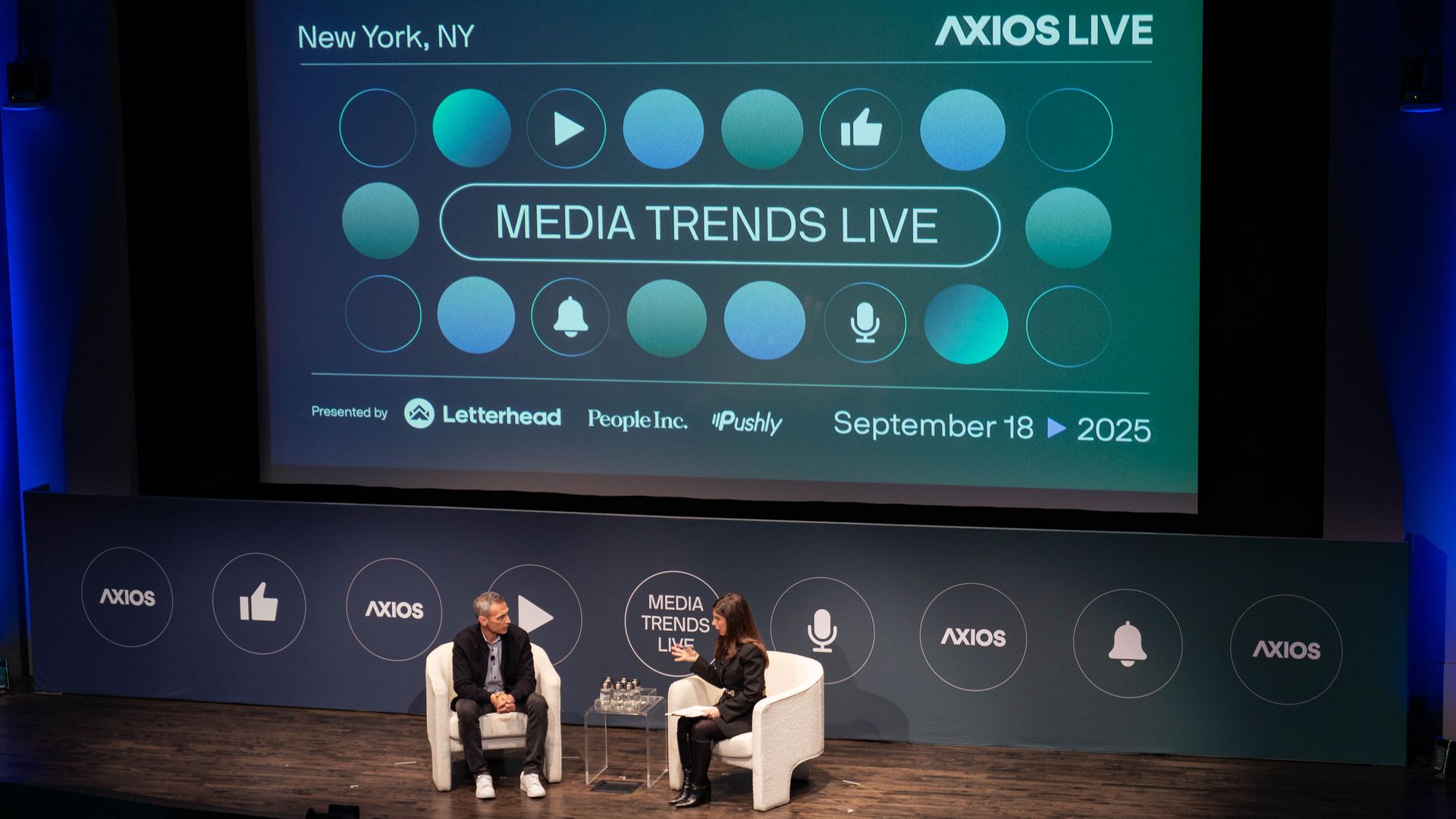 Two people seated on stage in front of a large screen displaying "MEDIA TRENDS LIVE," AXIOS LIVE, and event details for September 18, 2025 in New York, NY with blue and black colors.