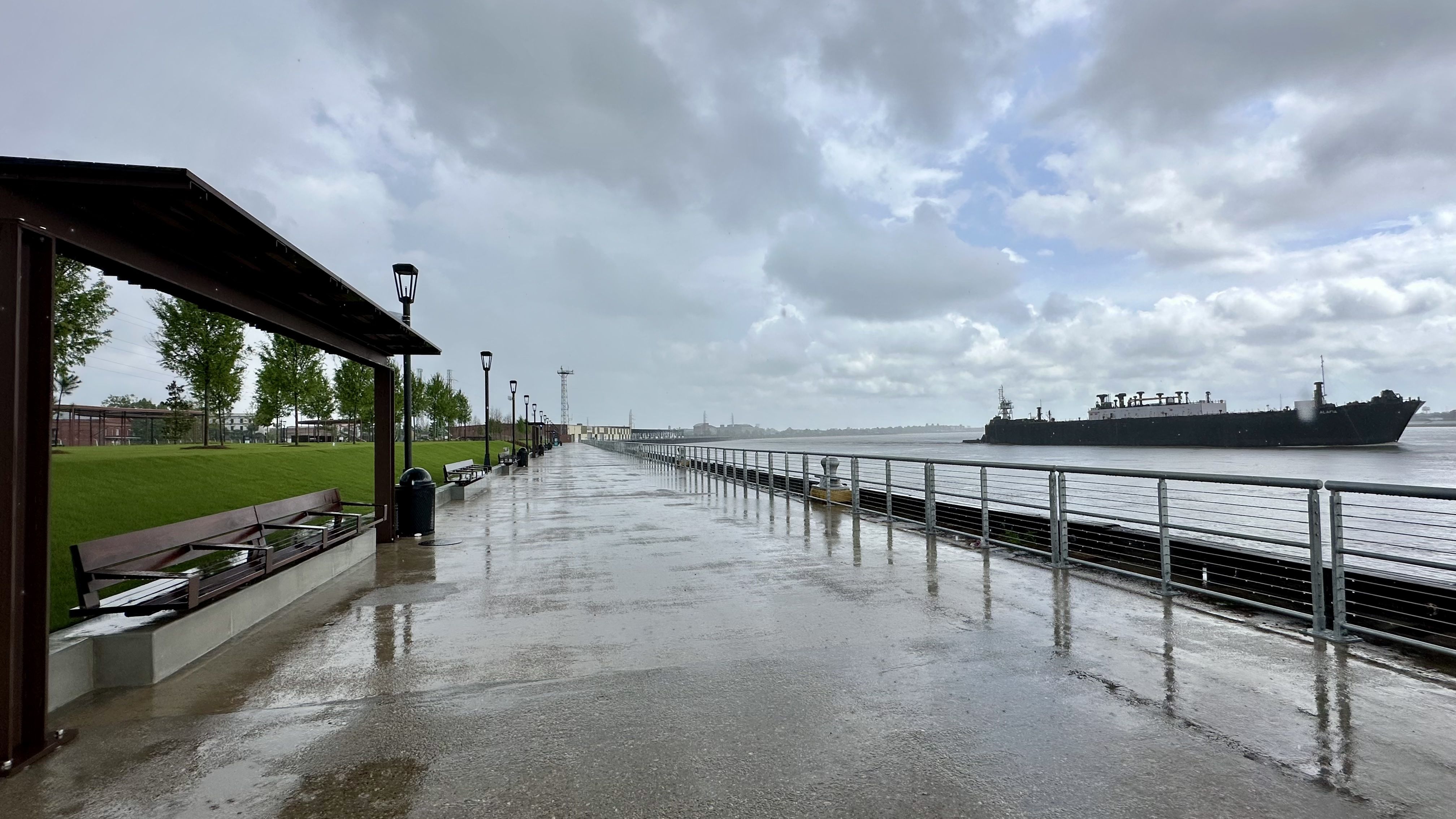 Wide riverside promenade on a rainy day: a wet concrete walkway with railing along the water, lamps, and benches, a green park with trees on the left, as a large cargo ship passes under cloudy skies.