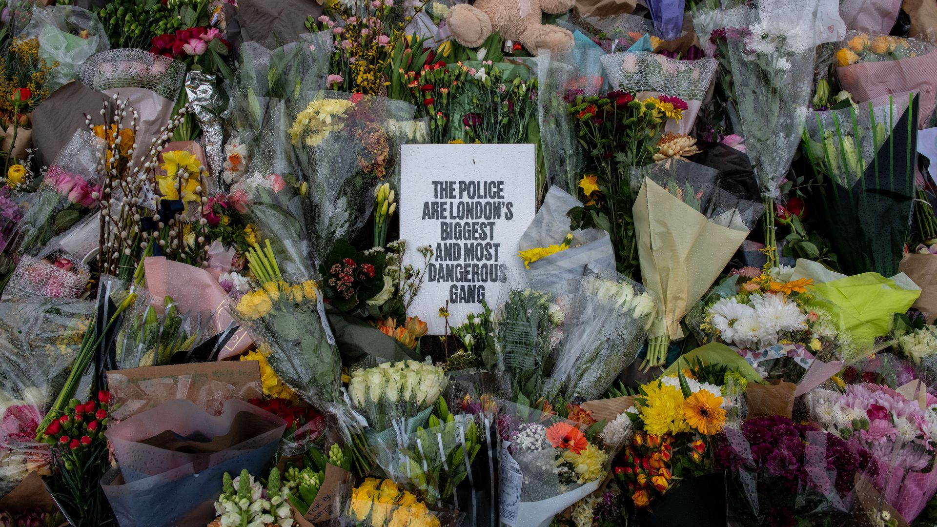 Picture of a floral tribute with flowers surrounding a sign that says "the police are London's biggest and most dangerous gang"
