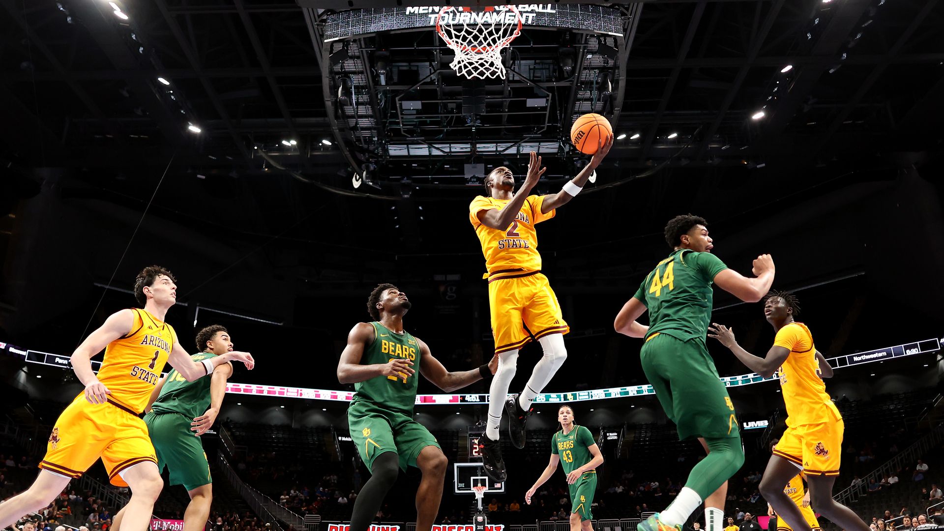 A basketball player in a yellow uniform jumps toward the rim for a layup while players in yellow and green uniforms surround him in a basketball arena.