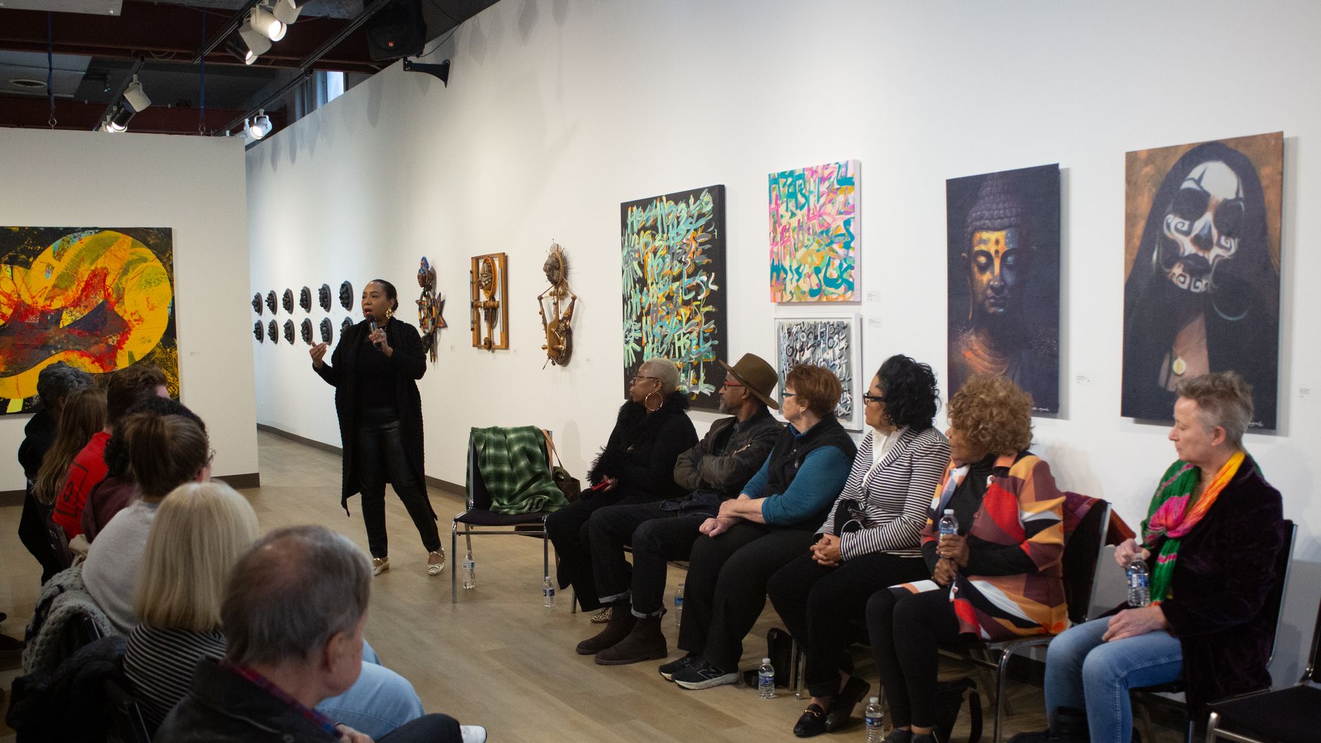 A person gives a gallery talk, in front of a white wall lined with colorful paintings and sculptures. The seated audience watches.