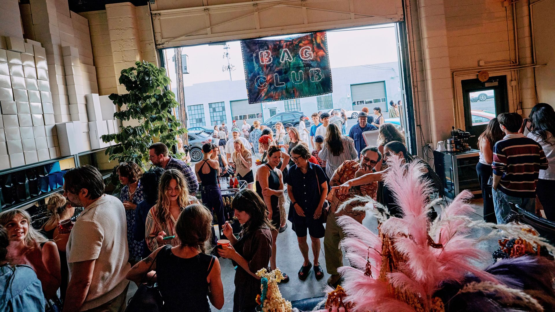 Inside a warehouse-style venue, a crowded party with people mingling around tables. A banner reading "BIG CLUB" hangs near the open garage door; pink feather decorations nearby.
