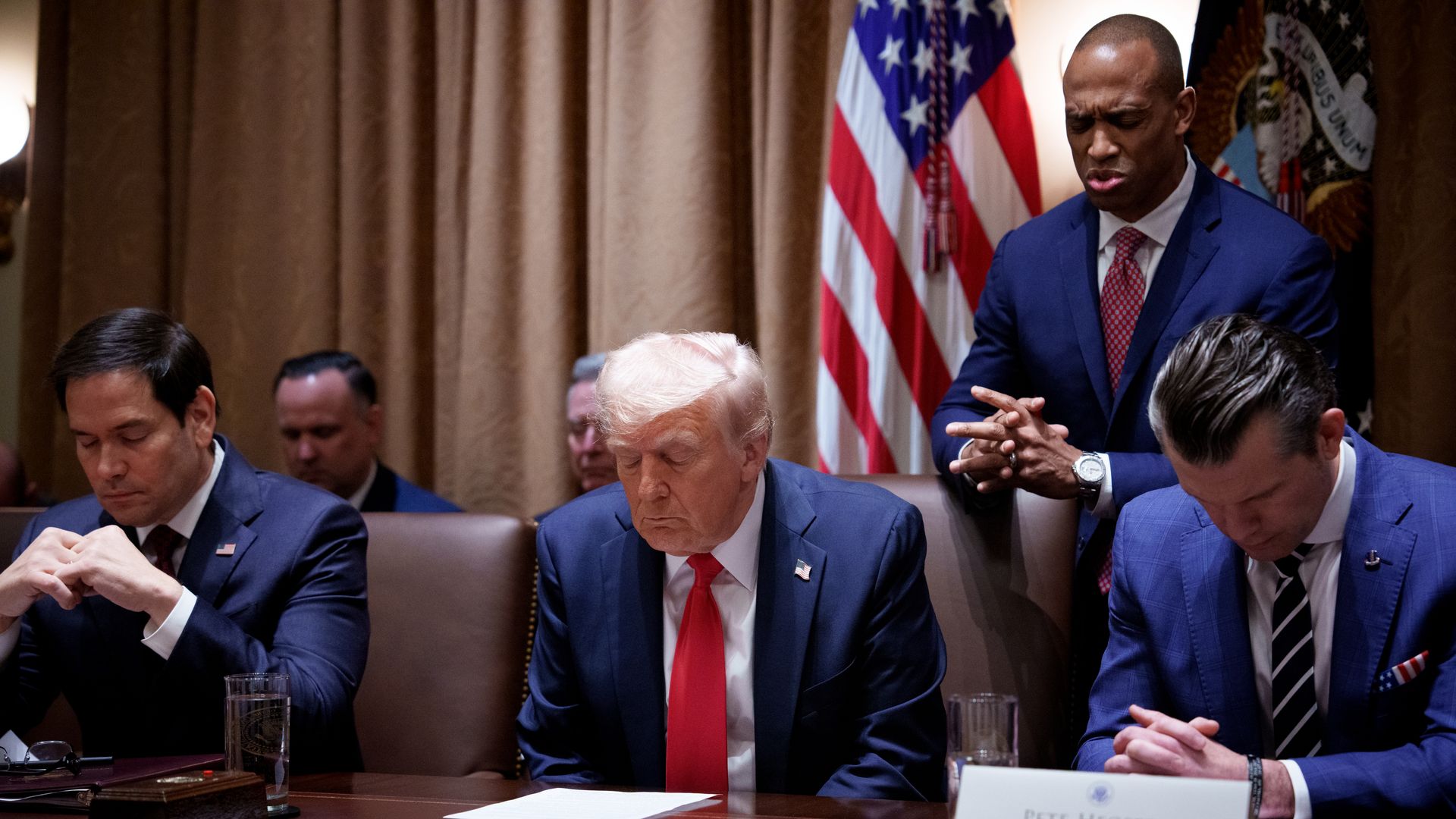President Trump bows his head in prayer during a Cabinet meeting whole wearing a white collared shirt, a blue suit jacket, a red tie and an American flag pin. Other members of his Cabinet sit around him, also wearing suits, while Scott Turner stands behind the president's chair.
