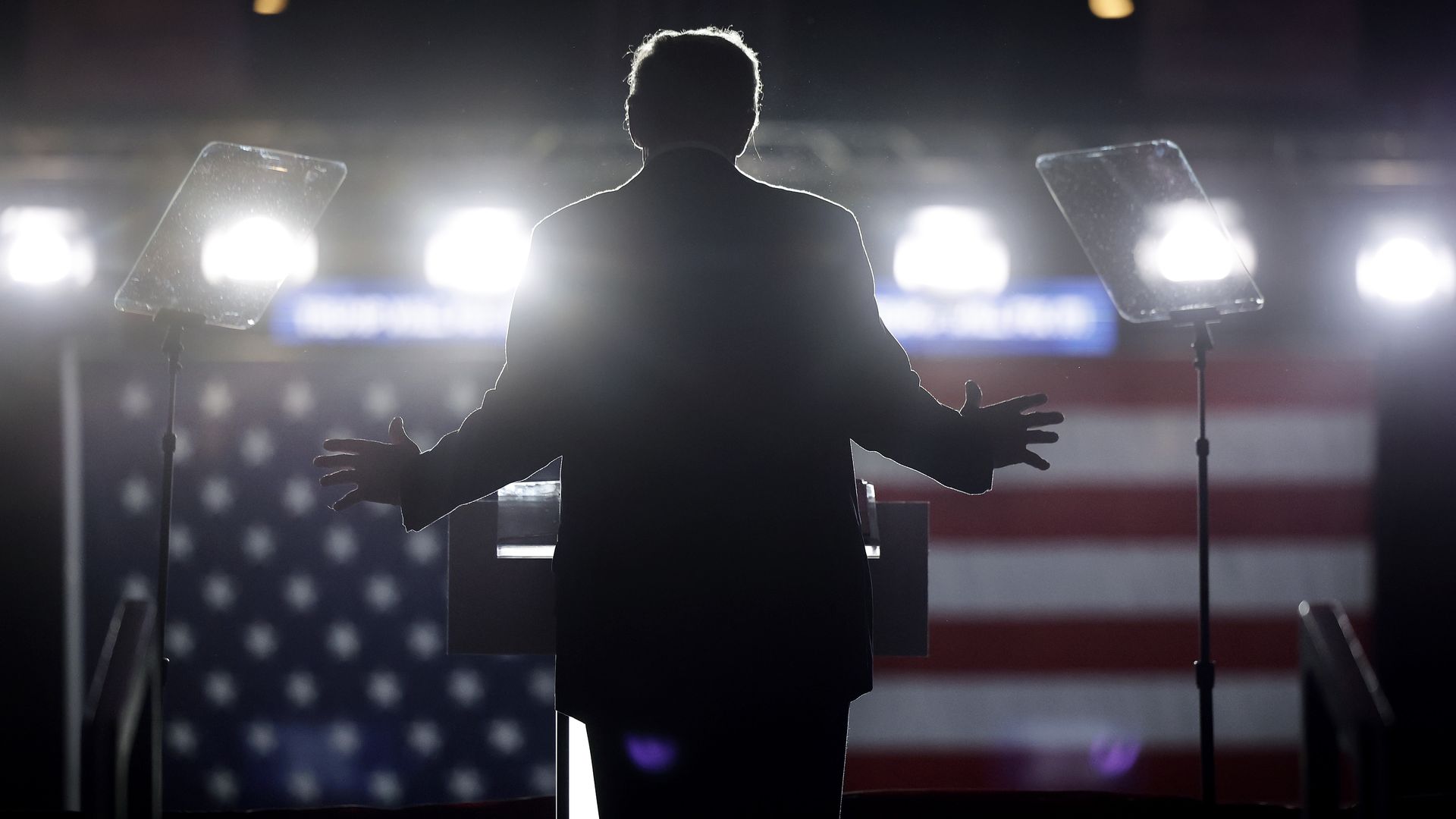 Donald Trump speaks on stage. He is backlit, with an American flag seen far away from him. He is waving his hands.