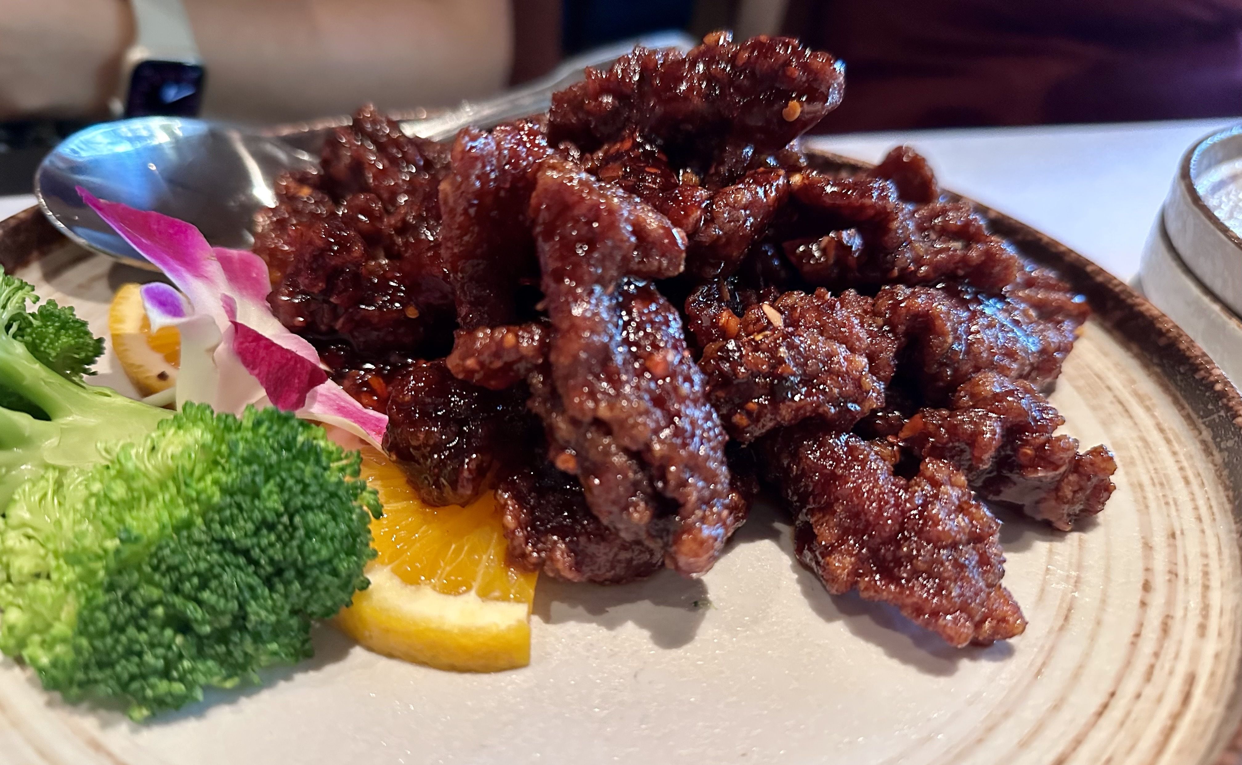 Plate of glazed, crispy dark brown beef strips garnished with fresh green broccoli, a slice of orange, and a purple and white orchid flower on a beige ceramic plate.