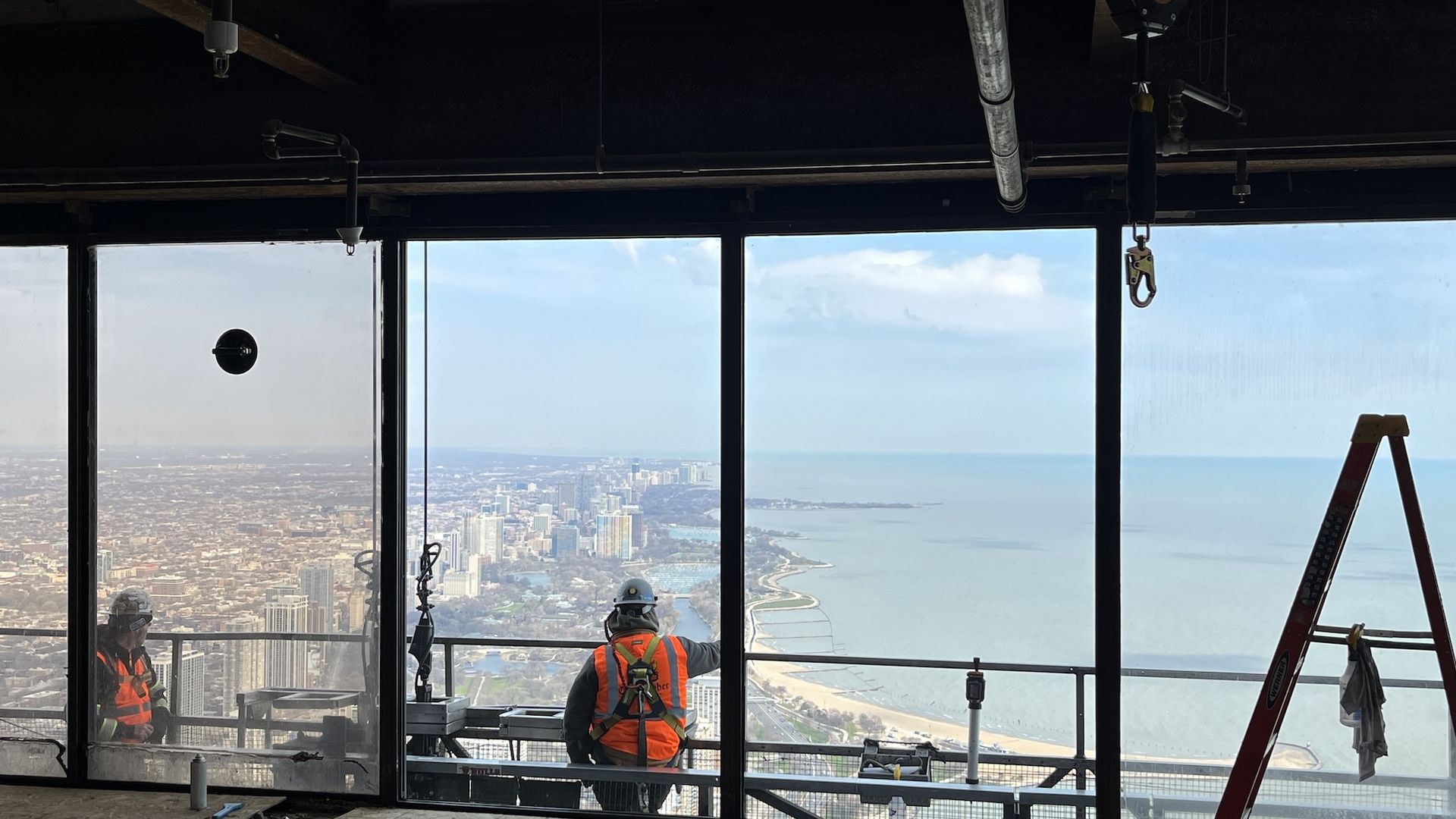 Construction site inside a high-rise with large glass windows, two workers in orange vests and hard hats overlook a city and shoreline, a red ladder on the right, exposed ducts above.