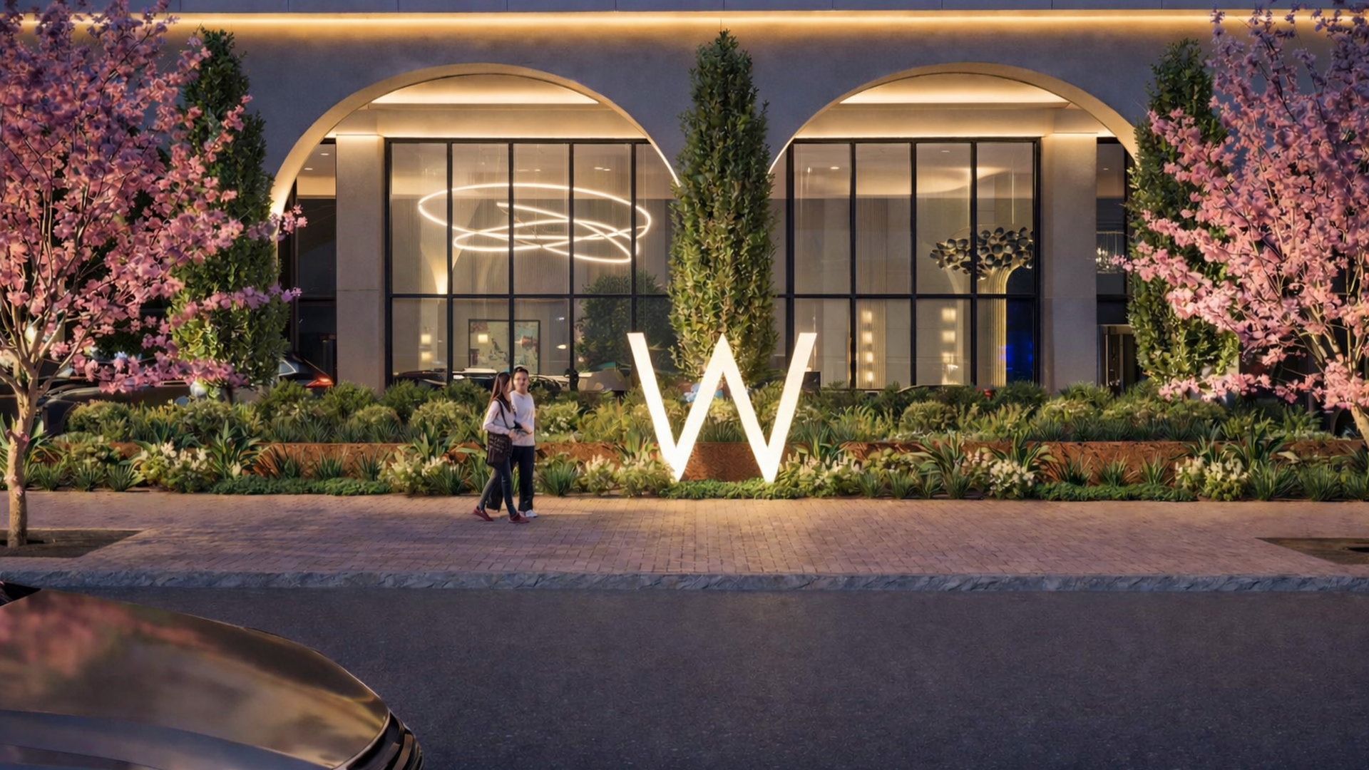 Modern storefront with a large illuminated white "W" sign front and center, flanked by pink blossoms and green shrubs. Two people stand by the sign as warm light glows through arched windows.