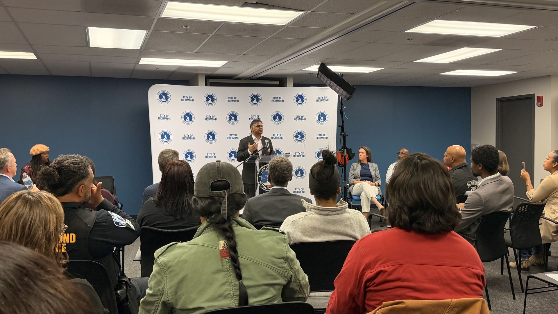 Man in a suit speaks at a podium during a City of Richmond event; a backdrop covered with circular logos and a blue wall, as a diverse audience listens in a conference room.