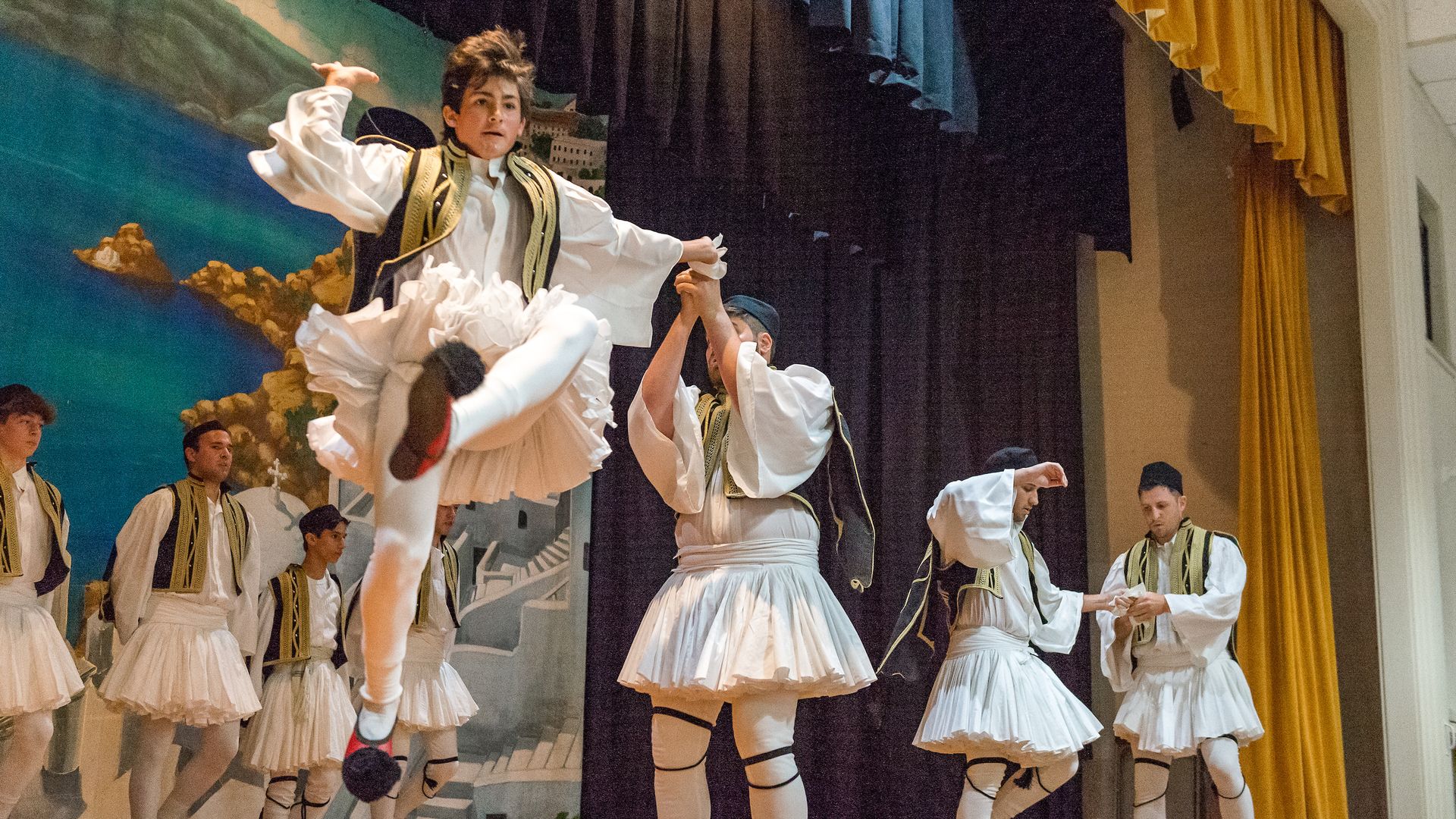Photo shows men dressed in traditional Greek clothes with tights and vests. They are dancing on a stage.