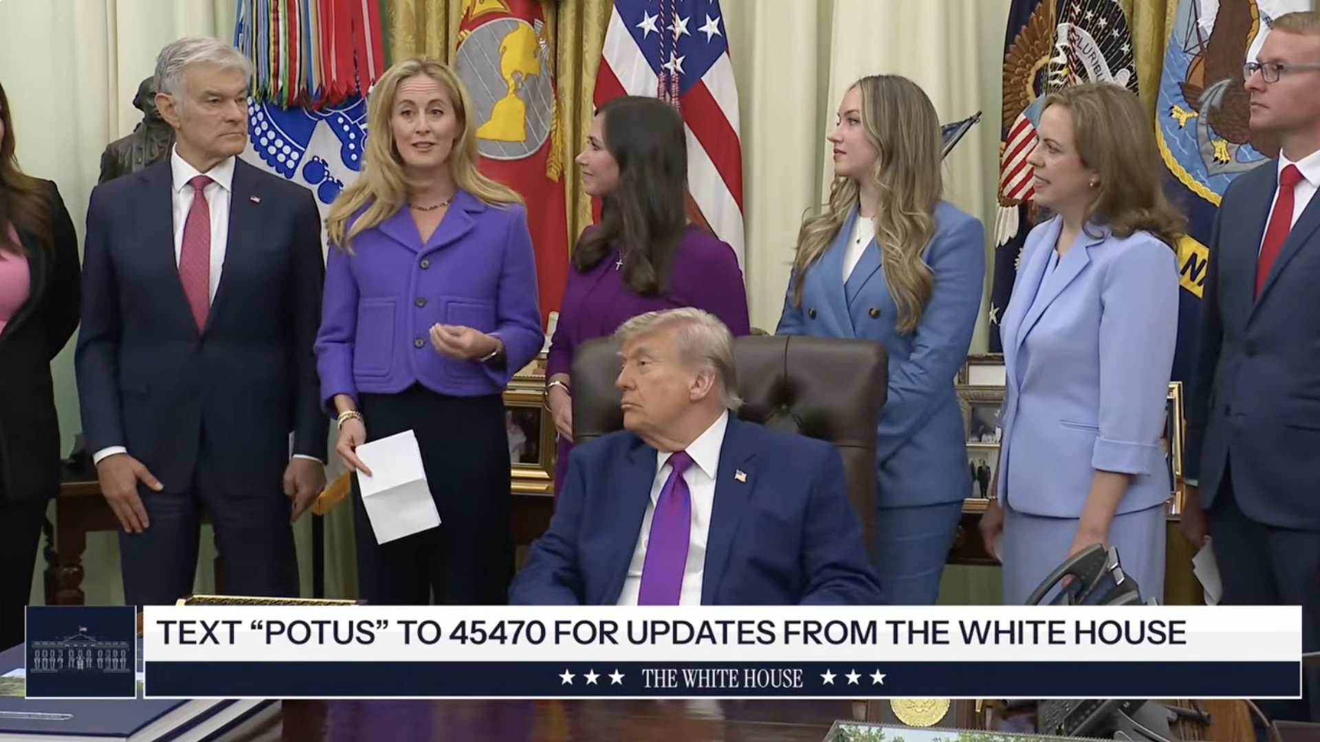 President Donald Trump sits at the Resolute Desk while a group of people stand behind him speaking in the Oval Office.
