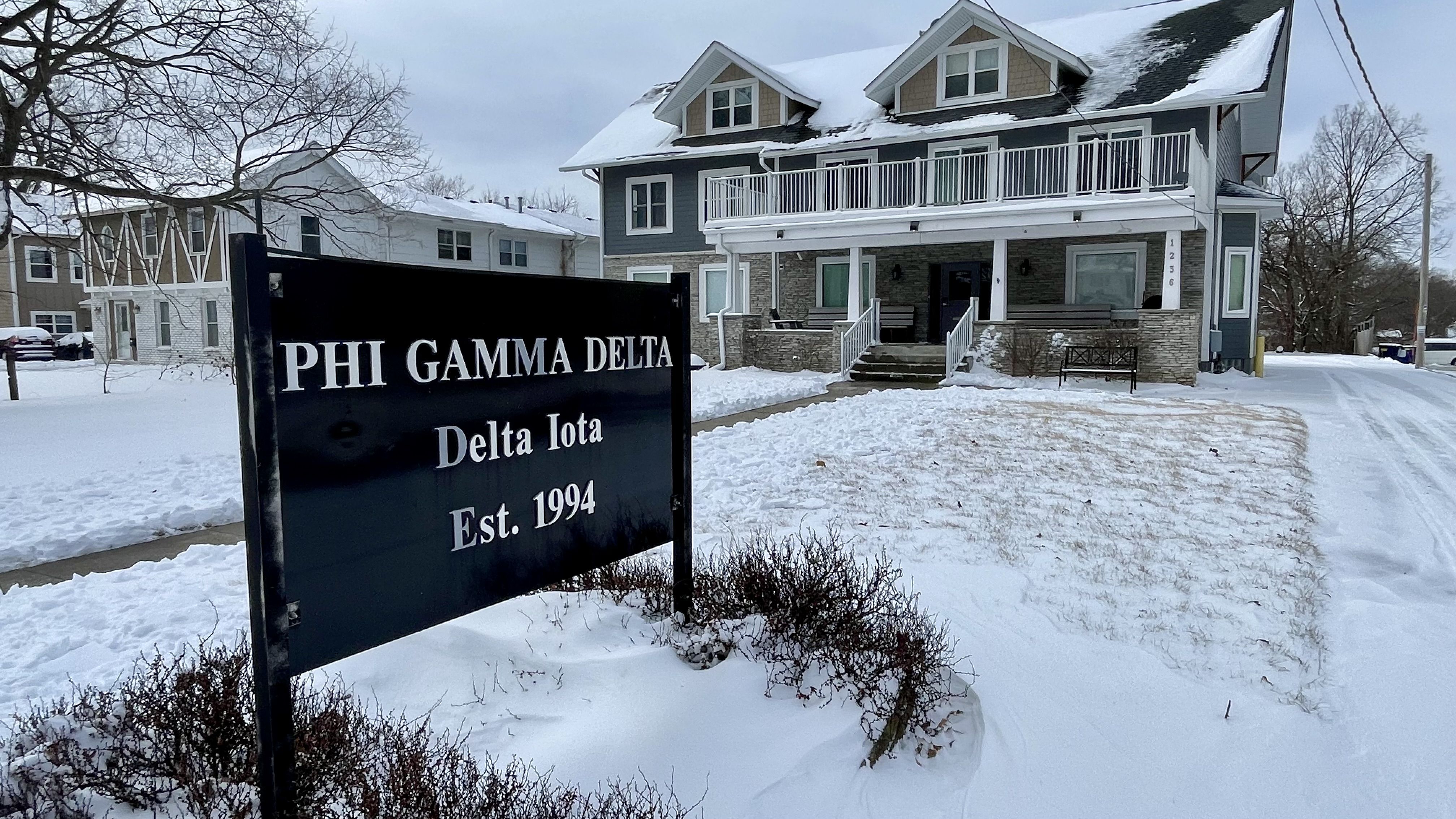 Snow-covered Phi Gamma Delta Delta Iota fraternity house with a dark blue sign showing establishment year 1994 in front of the two-story gray house on a winter day.