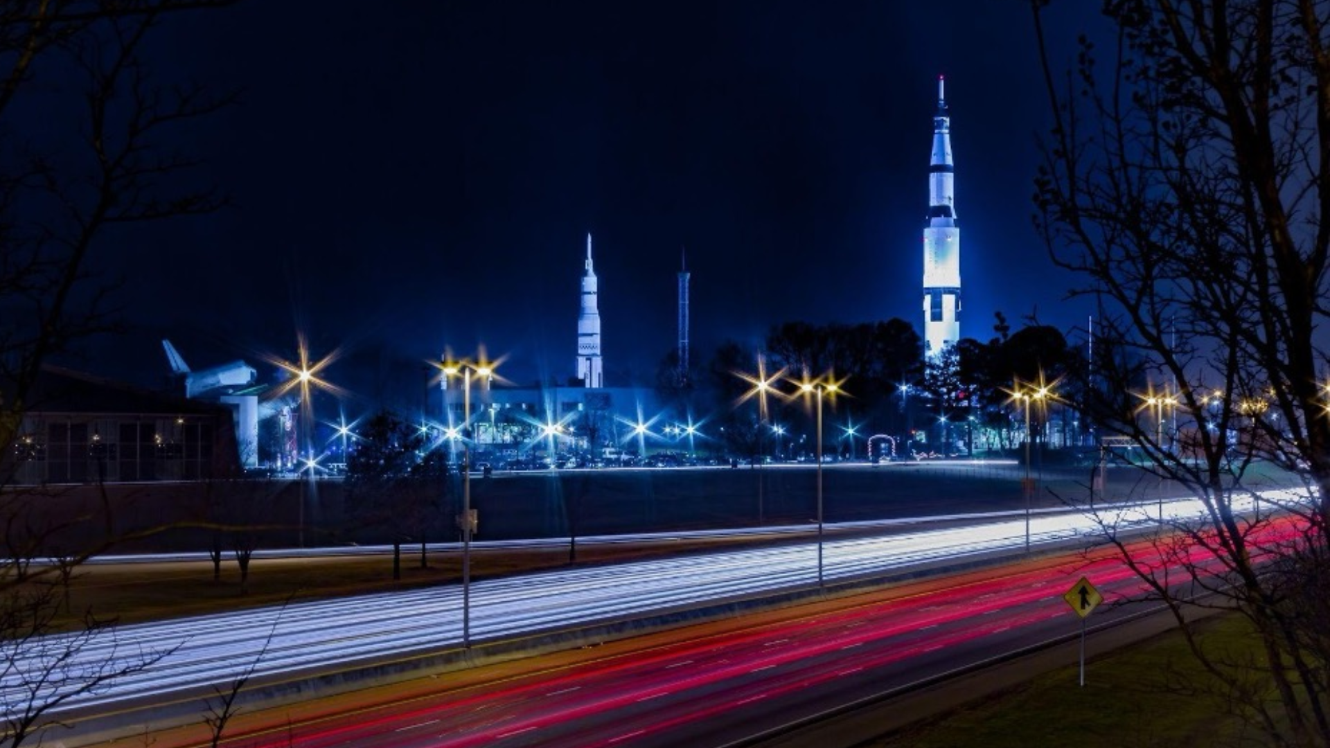 Night cityscape with a tall, illuminated tower and other buildings bathed in blue light; leafless trees frame the scene as blue streetlights and red/white light trails sweep across a highway.
