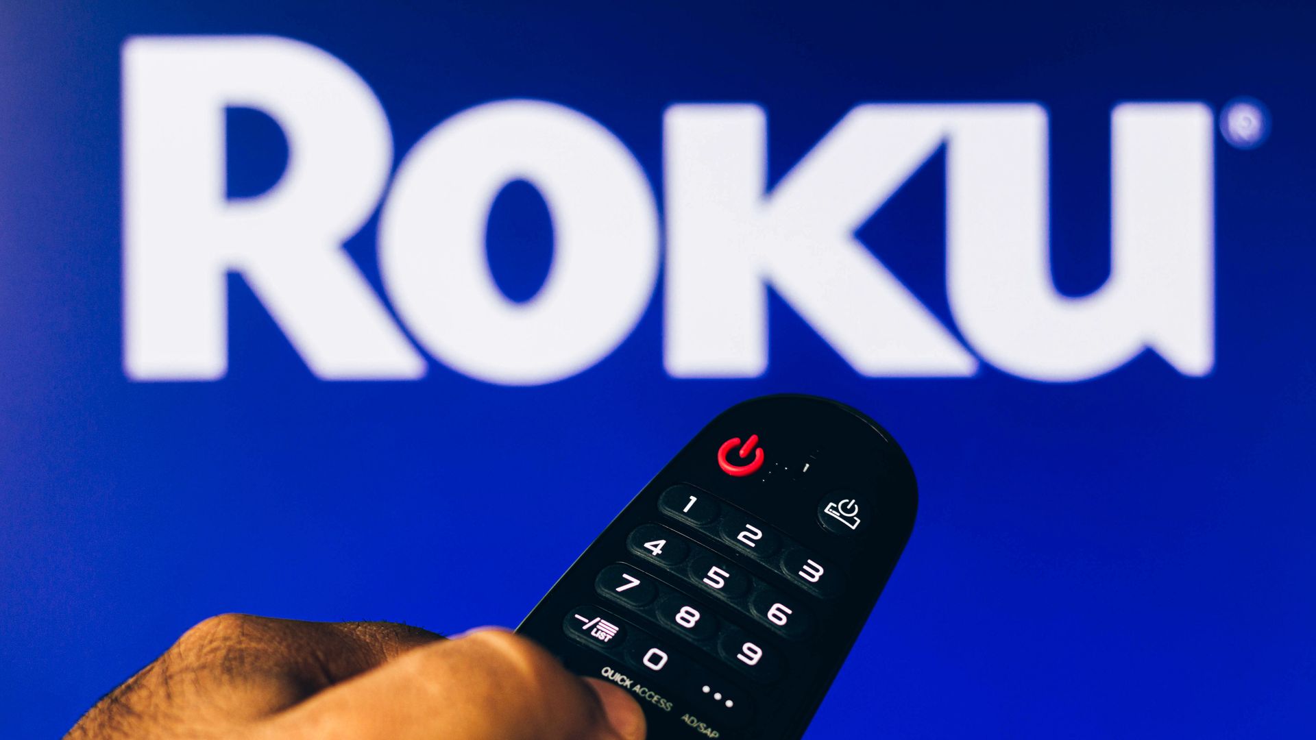 a hand holding a TV remote control points to a screen that displays the Roku logo