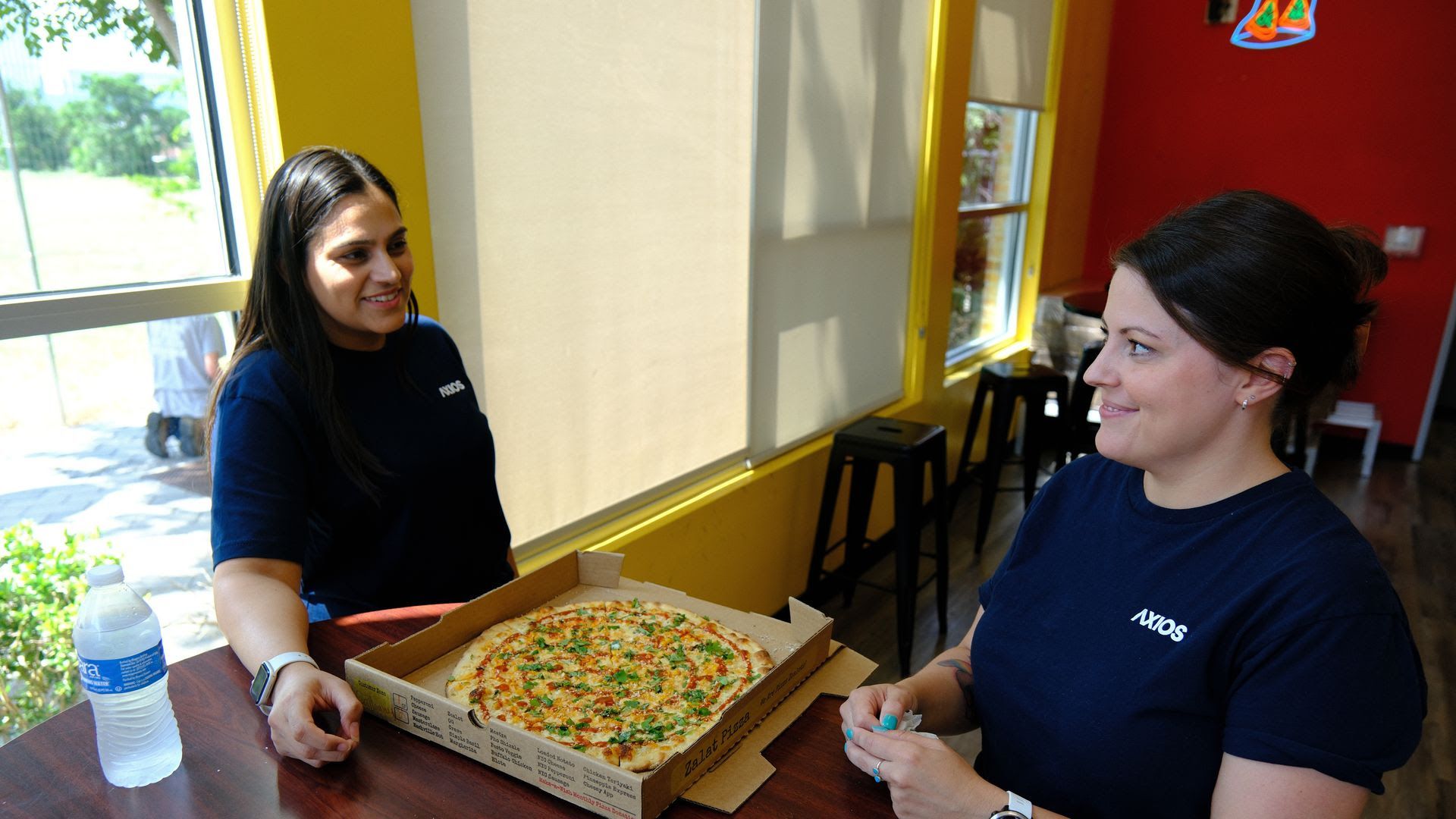 Two women standing at a table with a box of pizza on it