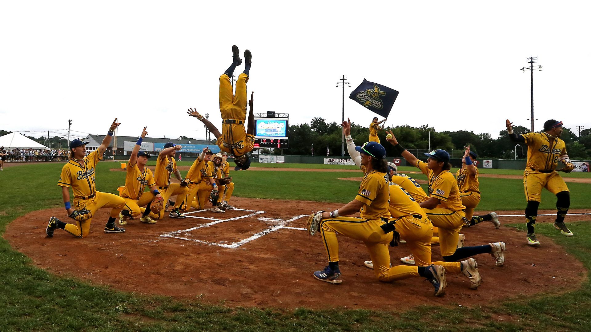 A baseball team in yellow uniforms kneel on either side of home plate as a player does a backflip