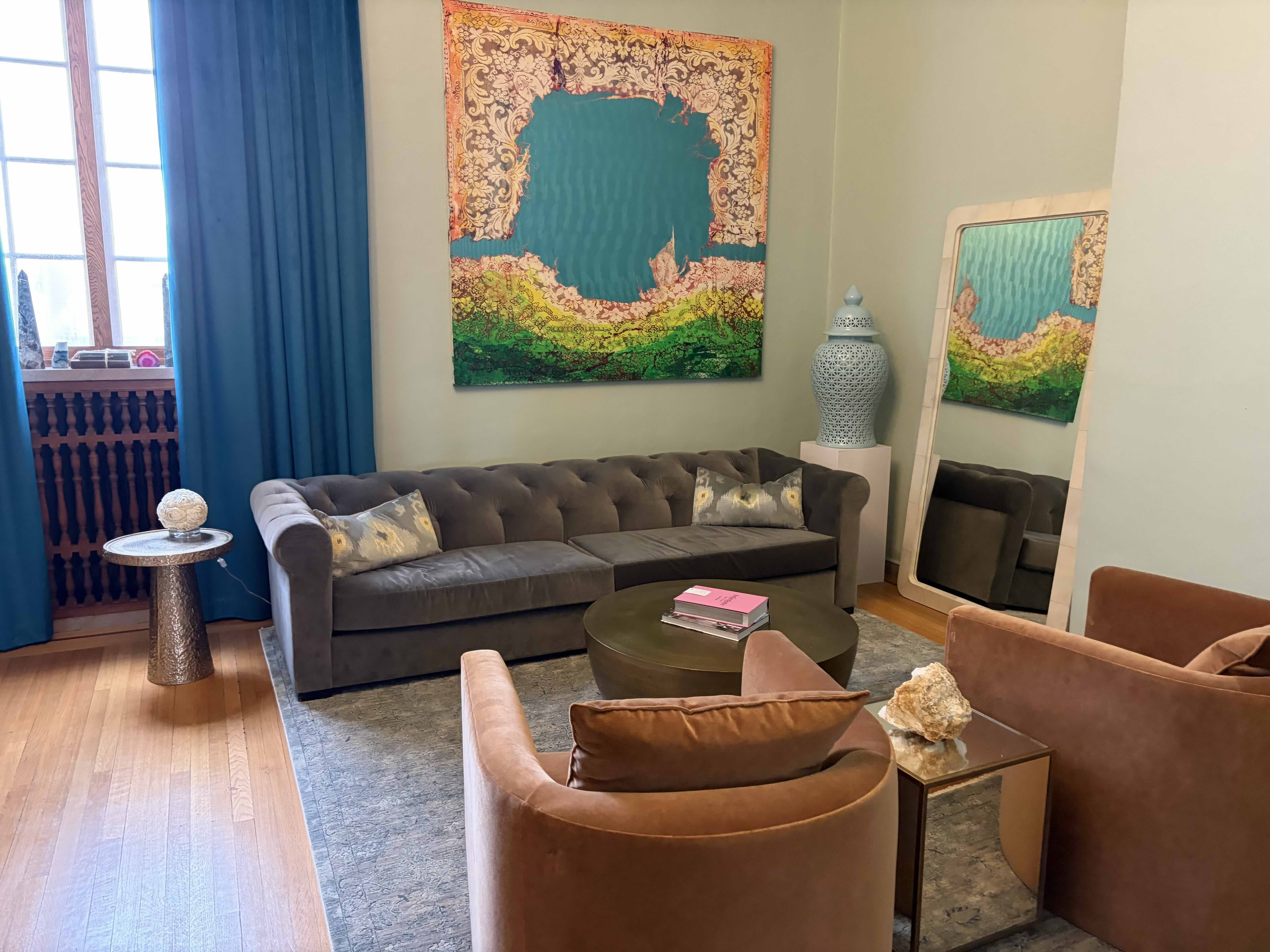 Cozy living room with a gray tufted sofa, teal curtains, and a bold painting featuring a turquoise center and ornate border. A blue lattice lamp sits on a pedestal beside a tall mirror reflecting art