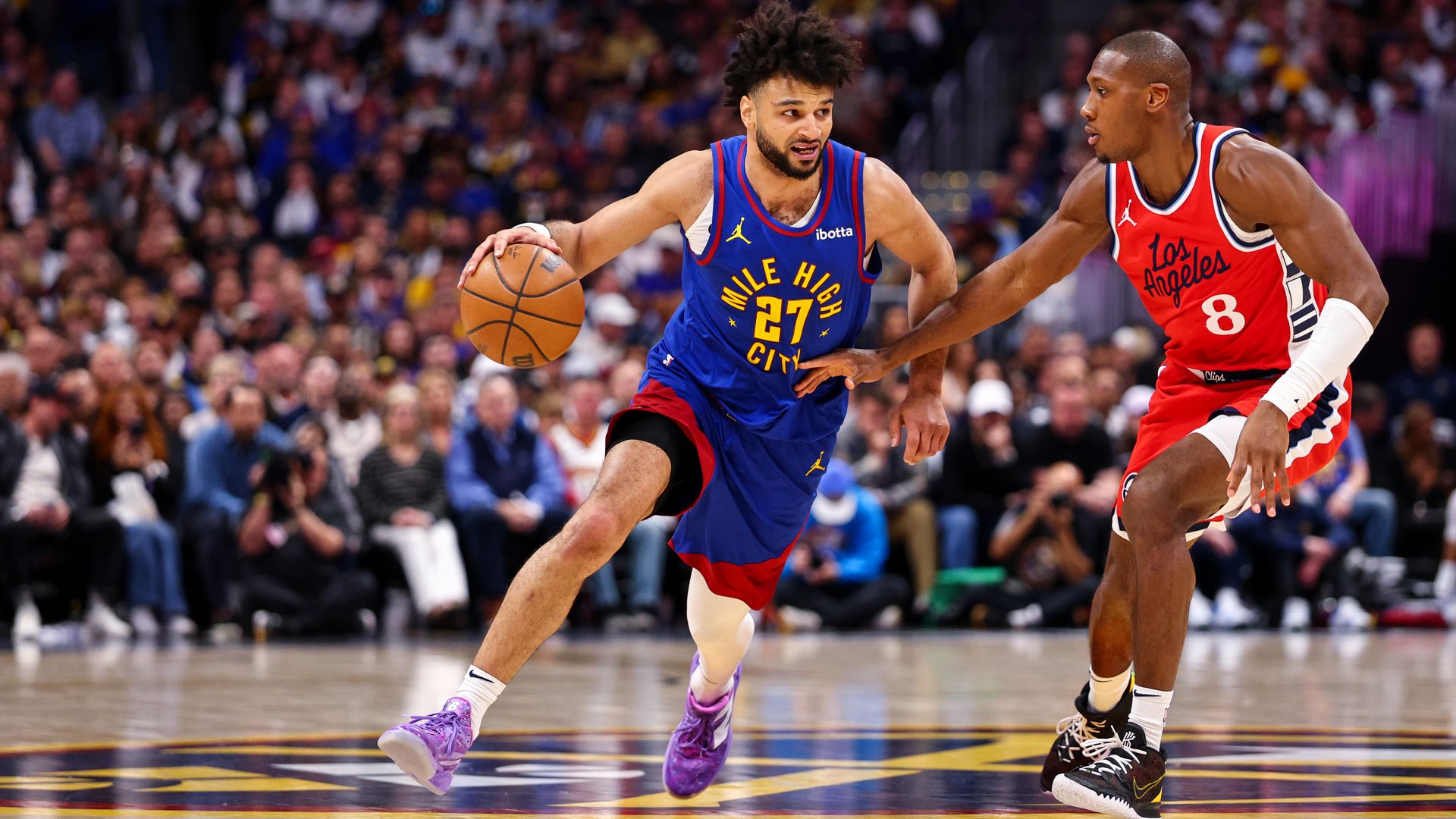 A man in a blue jersey handles a basketball while fending off an opposing player in a read jersey.