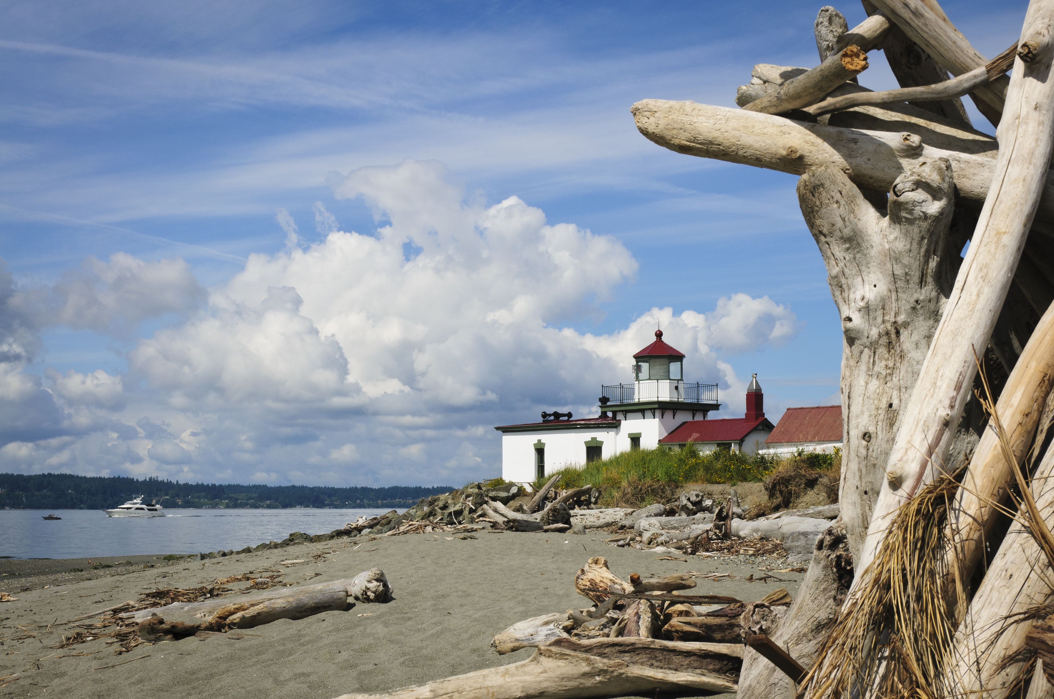 The West Point light house at Discovery Park is shown, with white paint and red roofing, on a sandy beach with logs strewn about, with a piece of driftwood in the foreground.