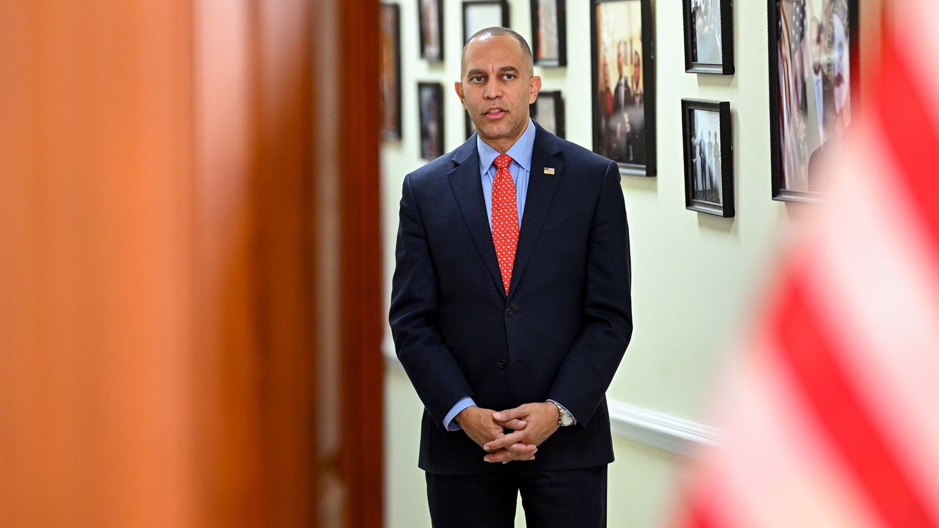 Hakeem Jeffries, wearing a blue suit and standing between a wooden doorway and a white hallway with photos on it.