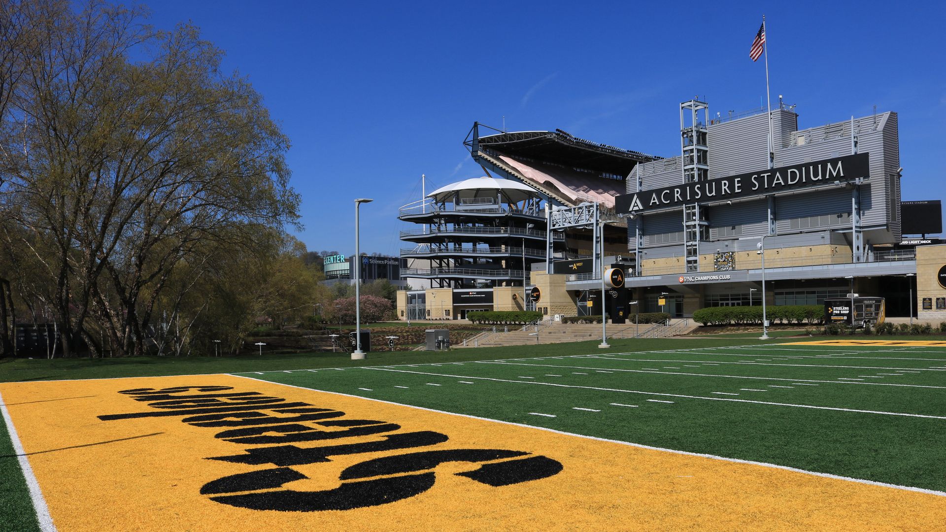 Exterior view of Acrisure Stadium on a clear day: blue sky, American flag atop the building, large stadium sign, green turf with white yard lines, and a yellow end zone with black lettering; trees to the left.