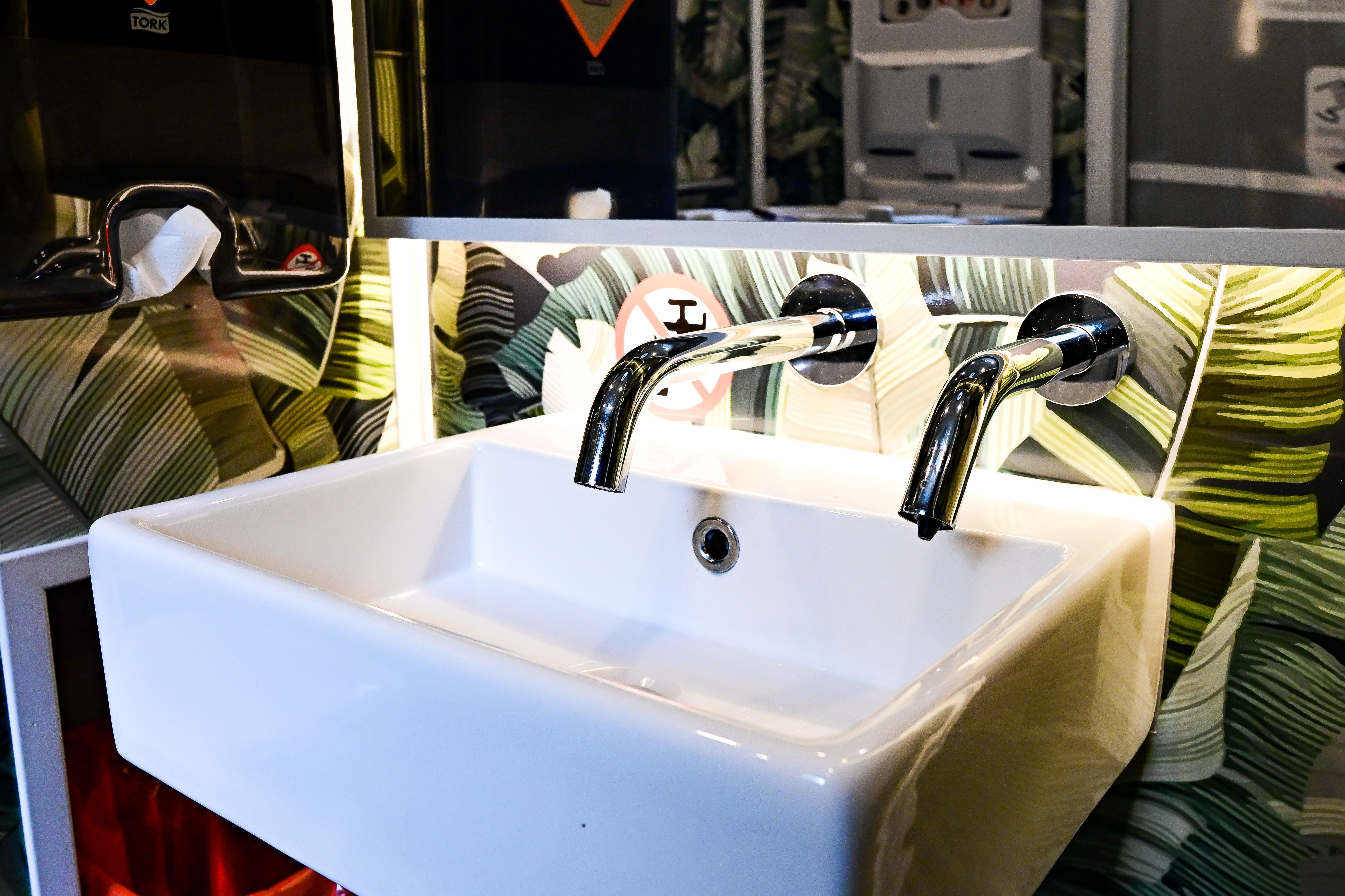 White rectangular wash basin with two curved chrome taps sits beneath a green tropical leaf-pattern wall; a tissue dispenser and dark cabinet are to the left, with metallic equipment overhead.