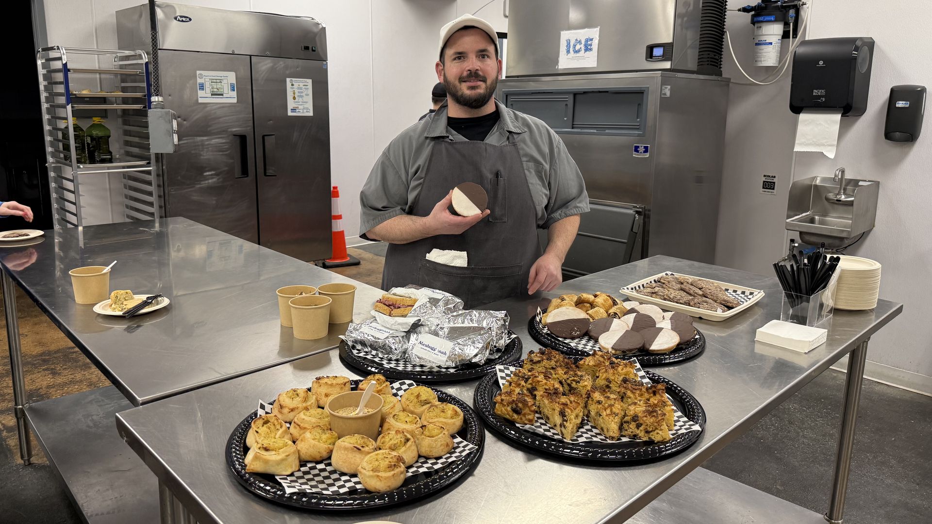 Man wearing apron and cap stands behind stainless steel table with trays of pastries, cookies, and drinks in a kitchen with ice machine and refrigerators.