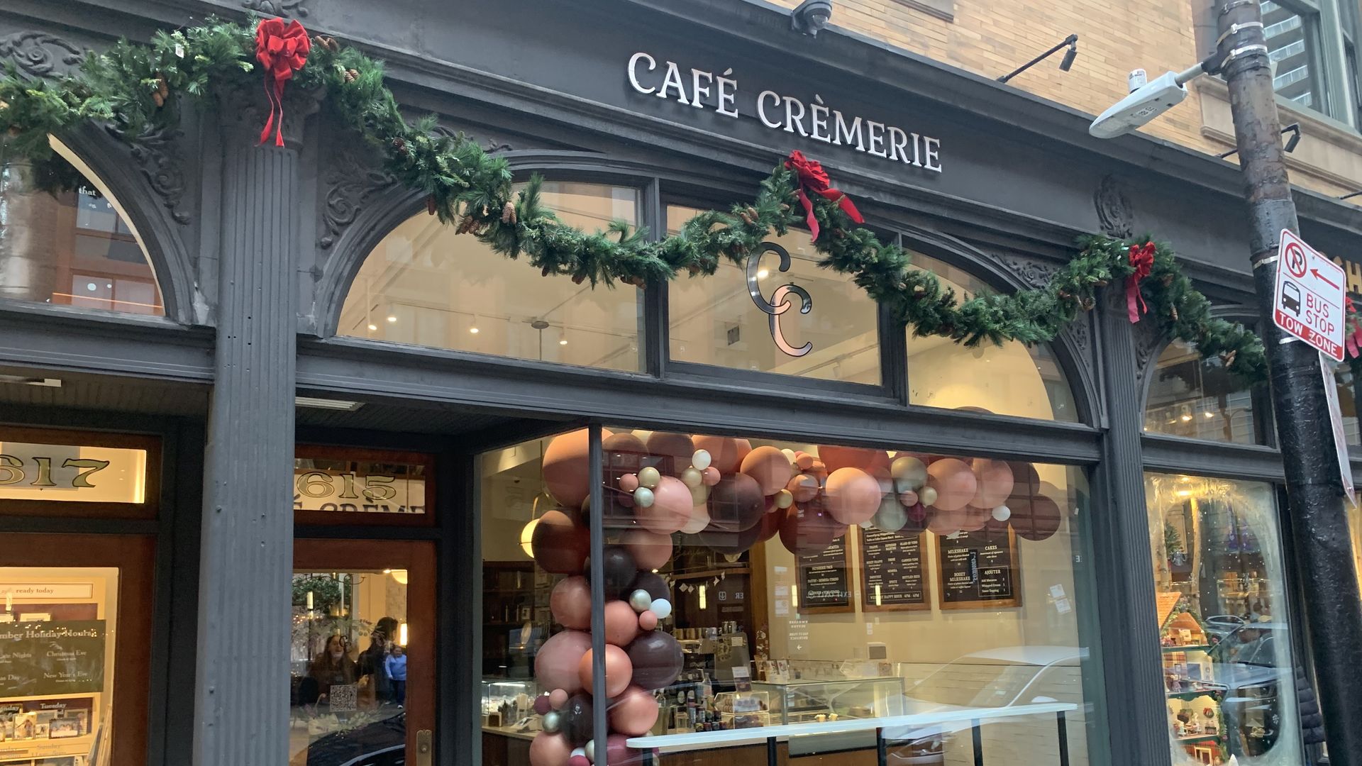 Exterior of a dark gray, ornate storefront; sign reads "CAFÉ CRÉMERIE". Festive garland with red bows decorates the facade, and large glass windows reveal balloons inside.