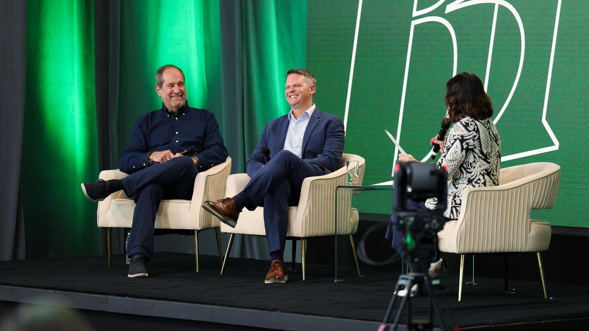 Two men are seated in chairs, smiling, while an interviewer asks a question.