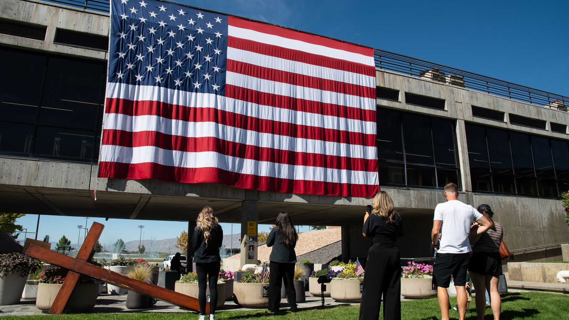 About five people stand in front of a fallen steel cross and a large American flag that hangs from a highway underpass.