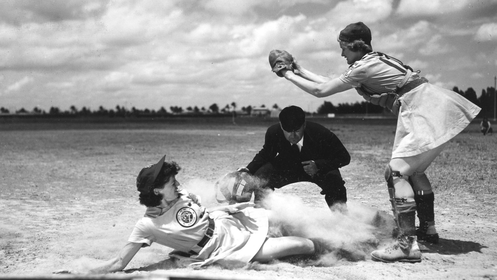 View of on-field action as All-American Girls Professional Baseball Leagues player Margaret 'Marge' Callaghan (later Maxwell, 1921 - 2019) (left), of the Fort Wayne Daisies, slides past umpire Norris 'Gadget' Ward (1907 - 1981) (center) while Callaghan's teammate Vivian 'Kelly' Kellogg (1922 - 2013)