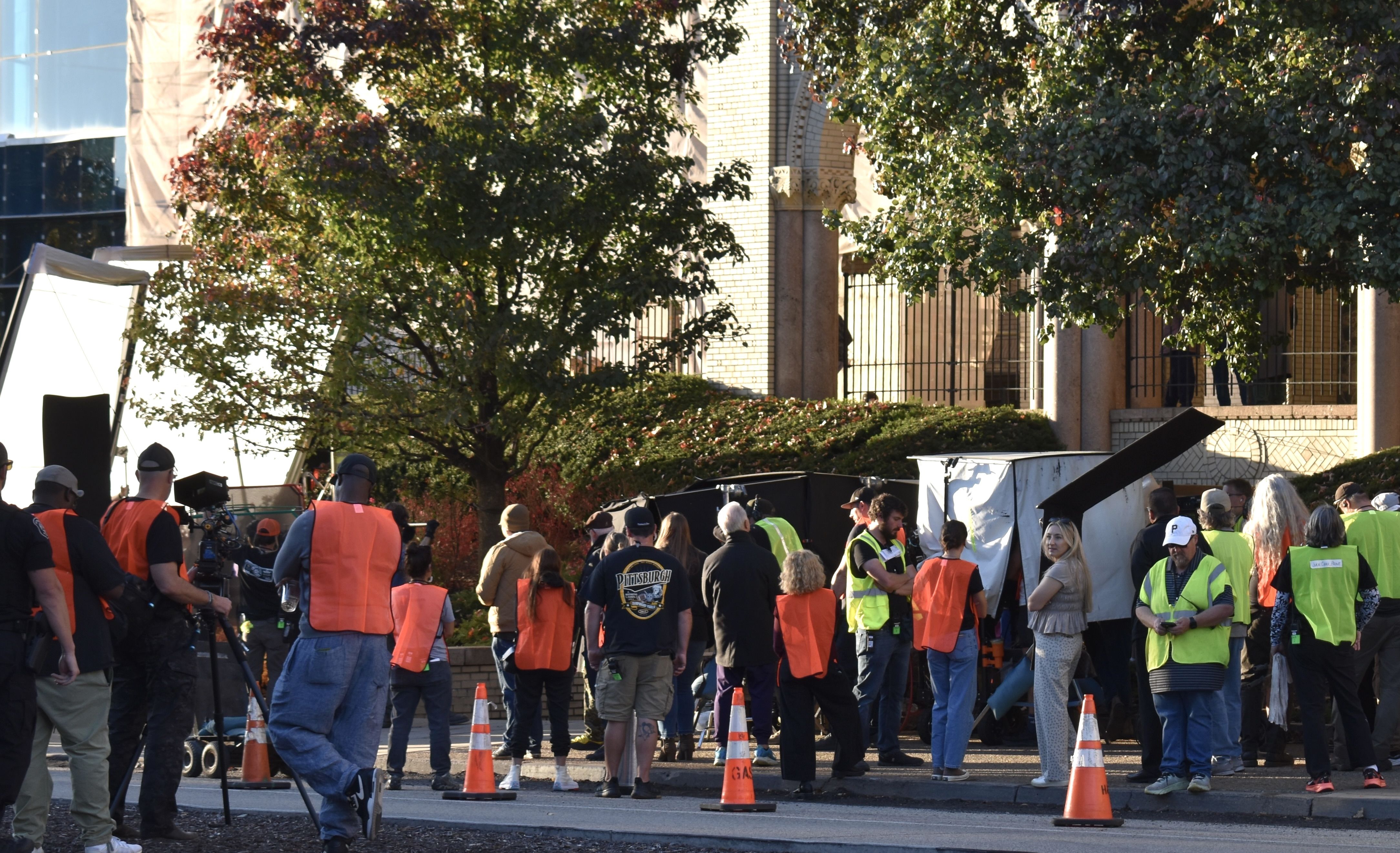 Wide crowd of people in orange and neon safety vests gathered on a street in front of a brick building with arches, orange cones, and trees; a few look like media crew and volunteers.