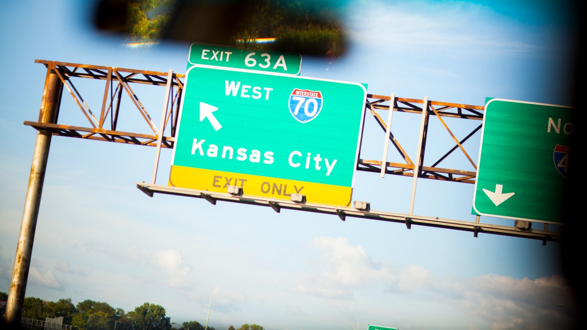 Photo shows a green interstate sign reading "WEST" and "I-70" and "Kansas City" and "EXIT ONLY" with an arrow pointing left from the perspective of inside a car with a rearview mirror in the foreground.