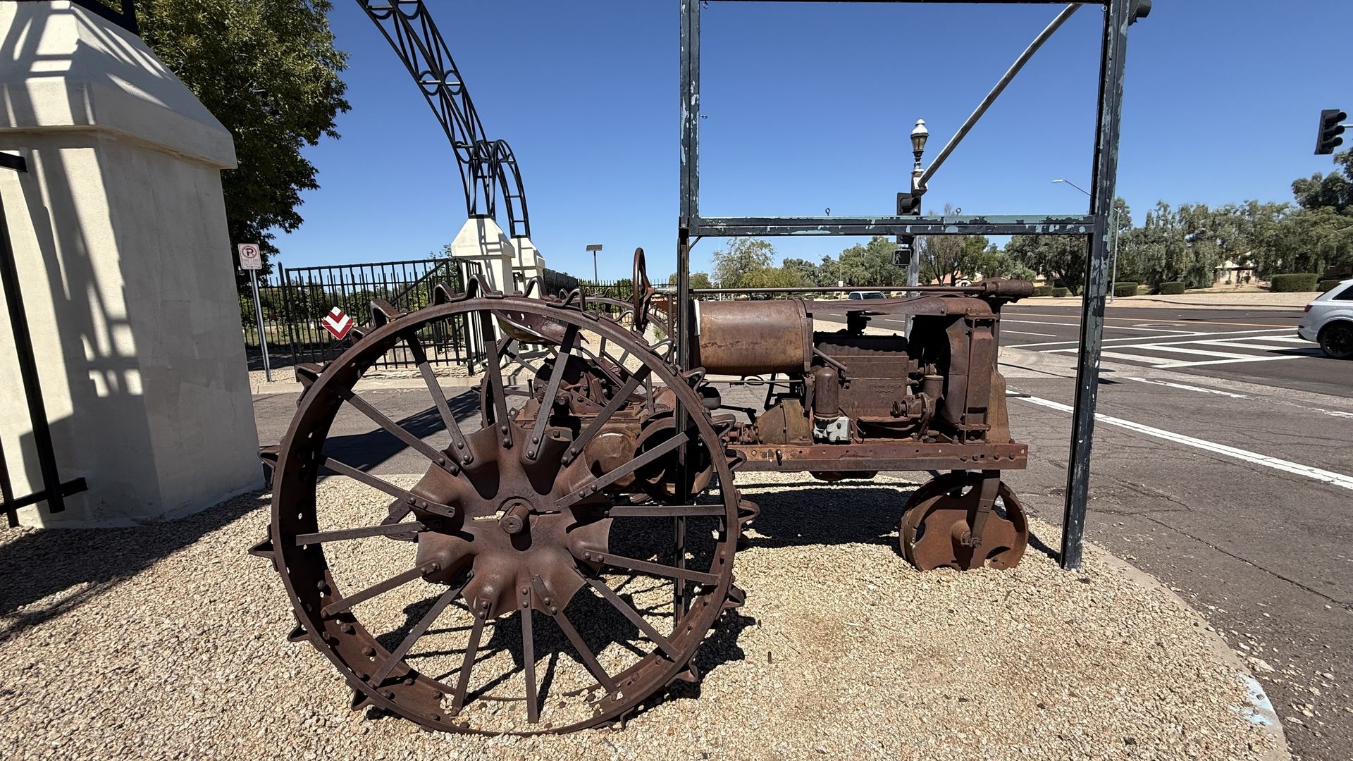 Rusty vintage tractor with large spiked wheels displayed outdoors on gravel near a street and a white concrete pillar.