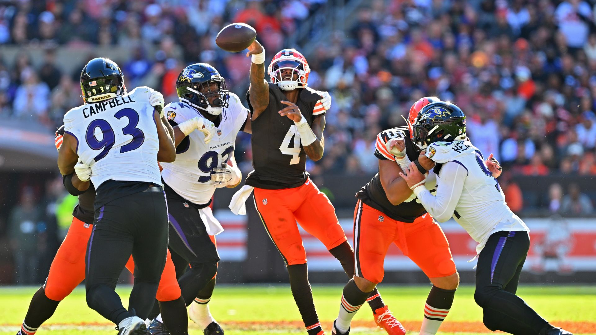An NFL moment: a Browns receiver leaps to catch a midair pass while Ravens defenders in white jerseys converge, on a sunlit field with a crowded stadium in the background.