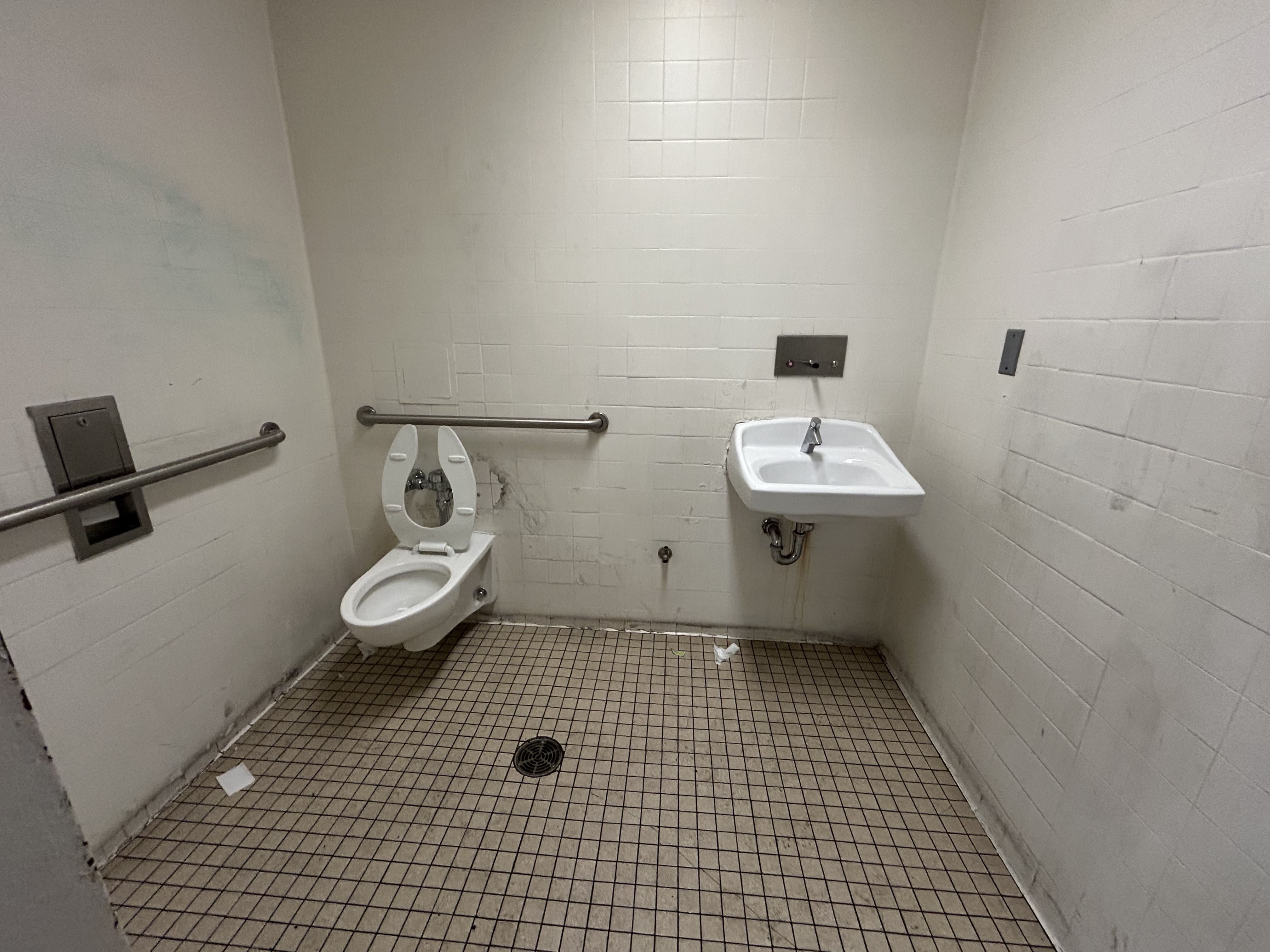 A dingy public restroom with white tile walls and a beige tiled floor. A wall-mounted sink on the right and a toilet with the seat up on the left; metal grab bars and a floor drain in the center.