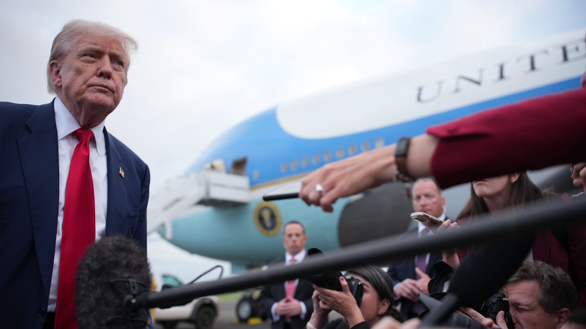 Man stands in red tie with plane in background and reporters in front