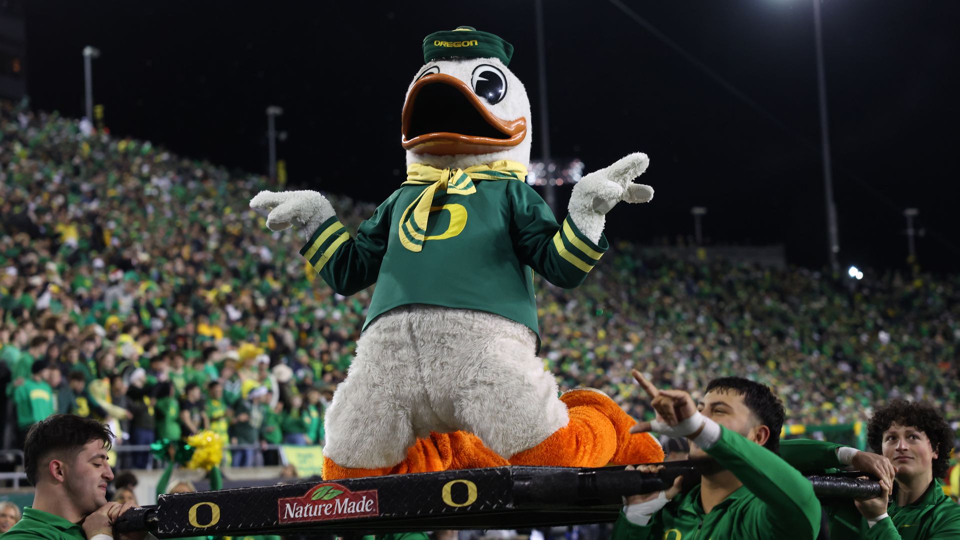 Night game in a packed stadium with green-and-yellow fans. A duck mascot in Oregon green uniform and cap stands on a railing, beak open, arms out, crowd cheering in the background.