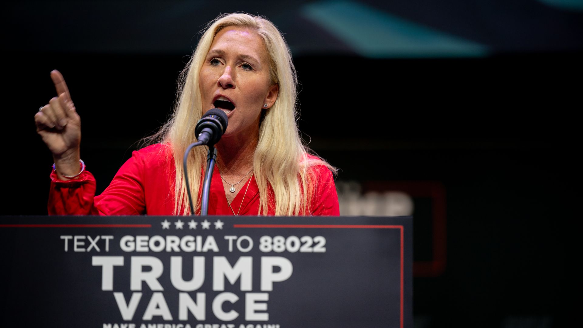 Rep. Marjorie Taylor Greene, wearing a red blouse, speaking at a Trump Vance podium in front of a dark backdrop.