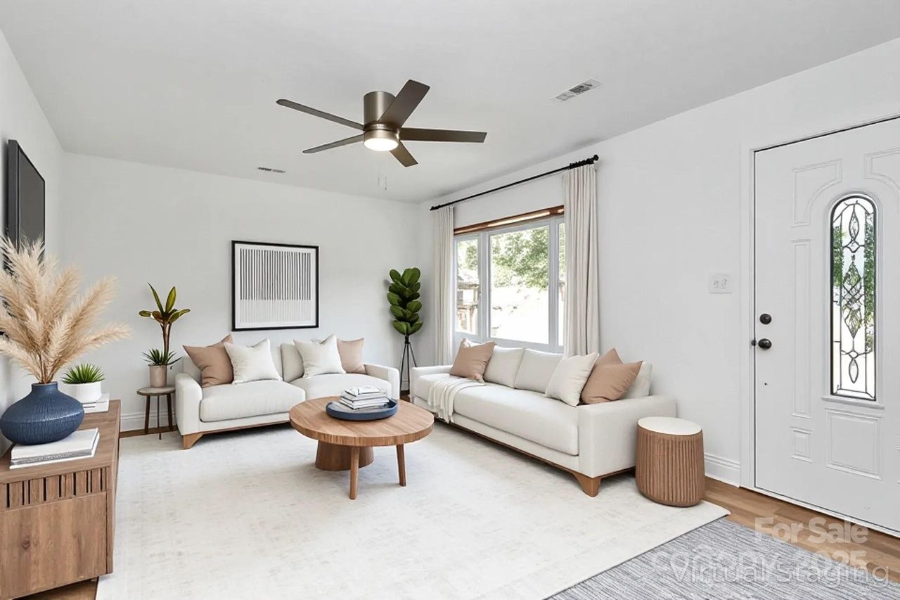 Bright living room with white walls, two beige sofas with brown and white cushions, round wooden coffee table, green plants, ceiling fan, large window with curtains, and white patterned rug.