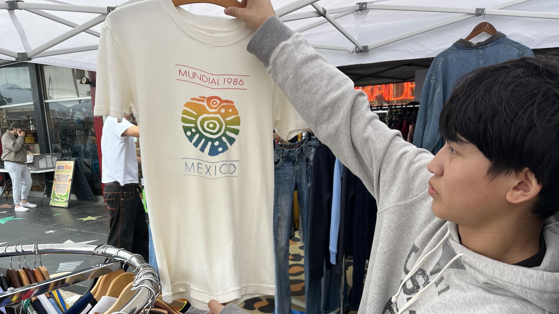 Young man in a gray hoodie holds up a cream T-shirt with a rainbow circular design and the text Mundial 1986 Mexico at a clothing stall under a tent.