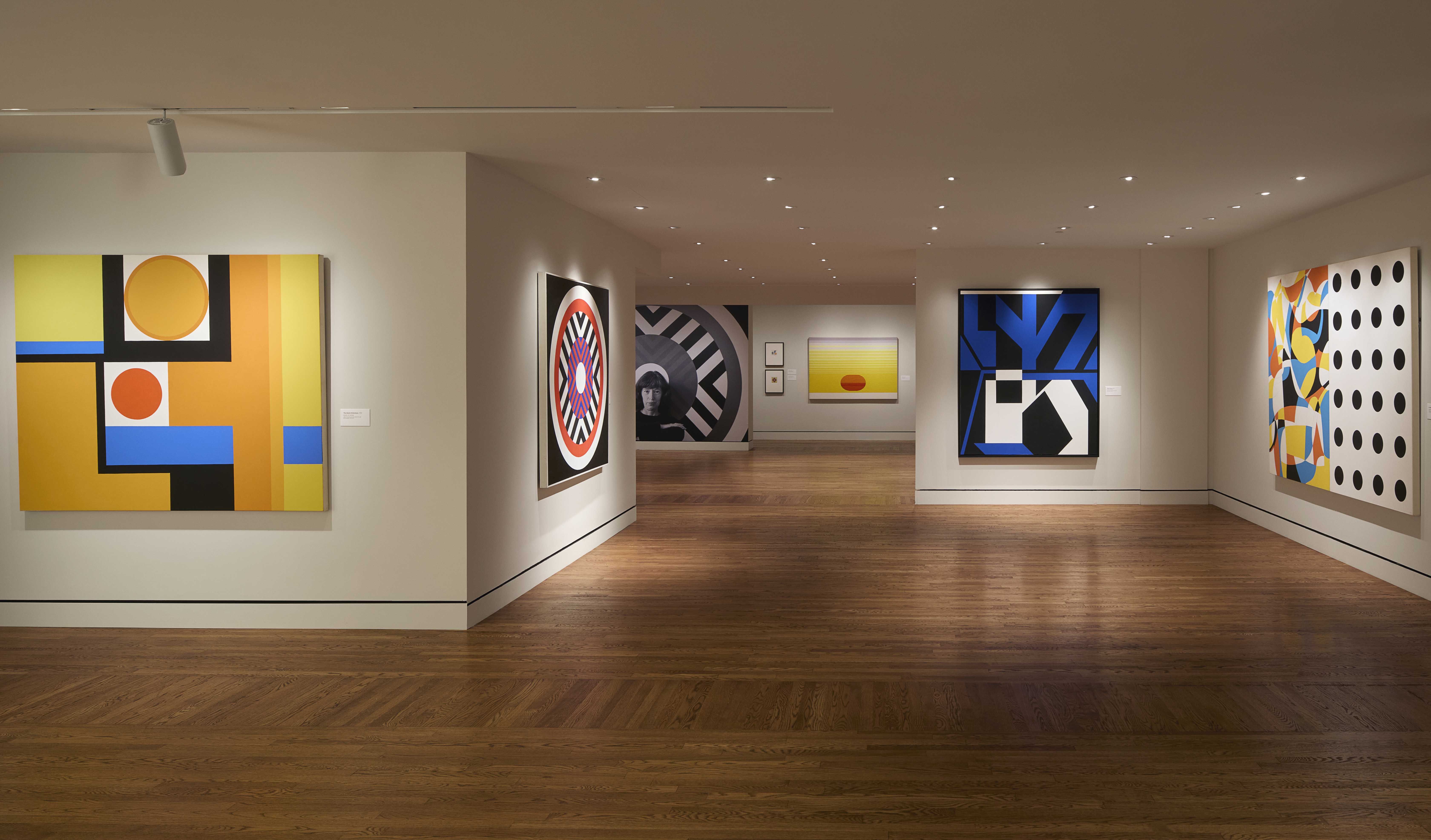 Art gallery room with wooden floor showing colorful geometric and abstract paintings in orange, yellow, blue, black, white, and multicolor on white walls under ceiling lights.