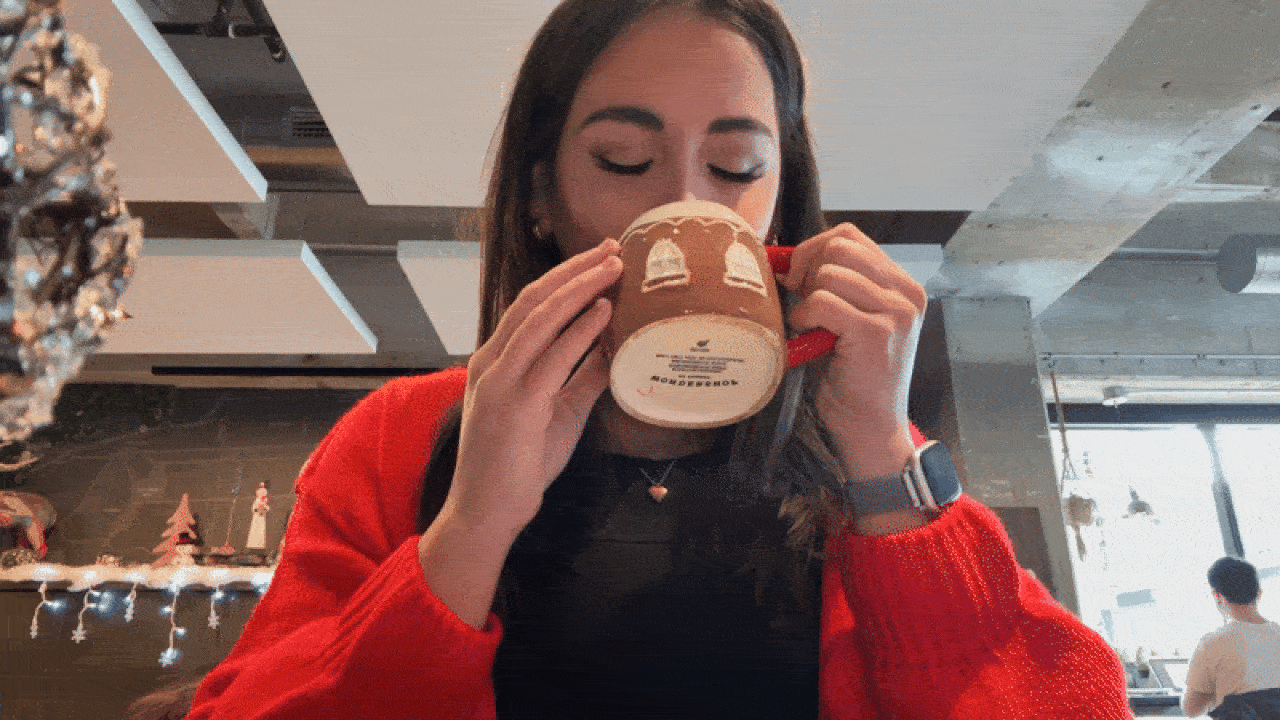 Woman in a red sweater sipping from a brown Christmas-themed mug with white bell designs, sitting in a cozy cafe decorated with holiday lights and ornaments.