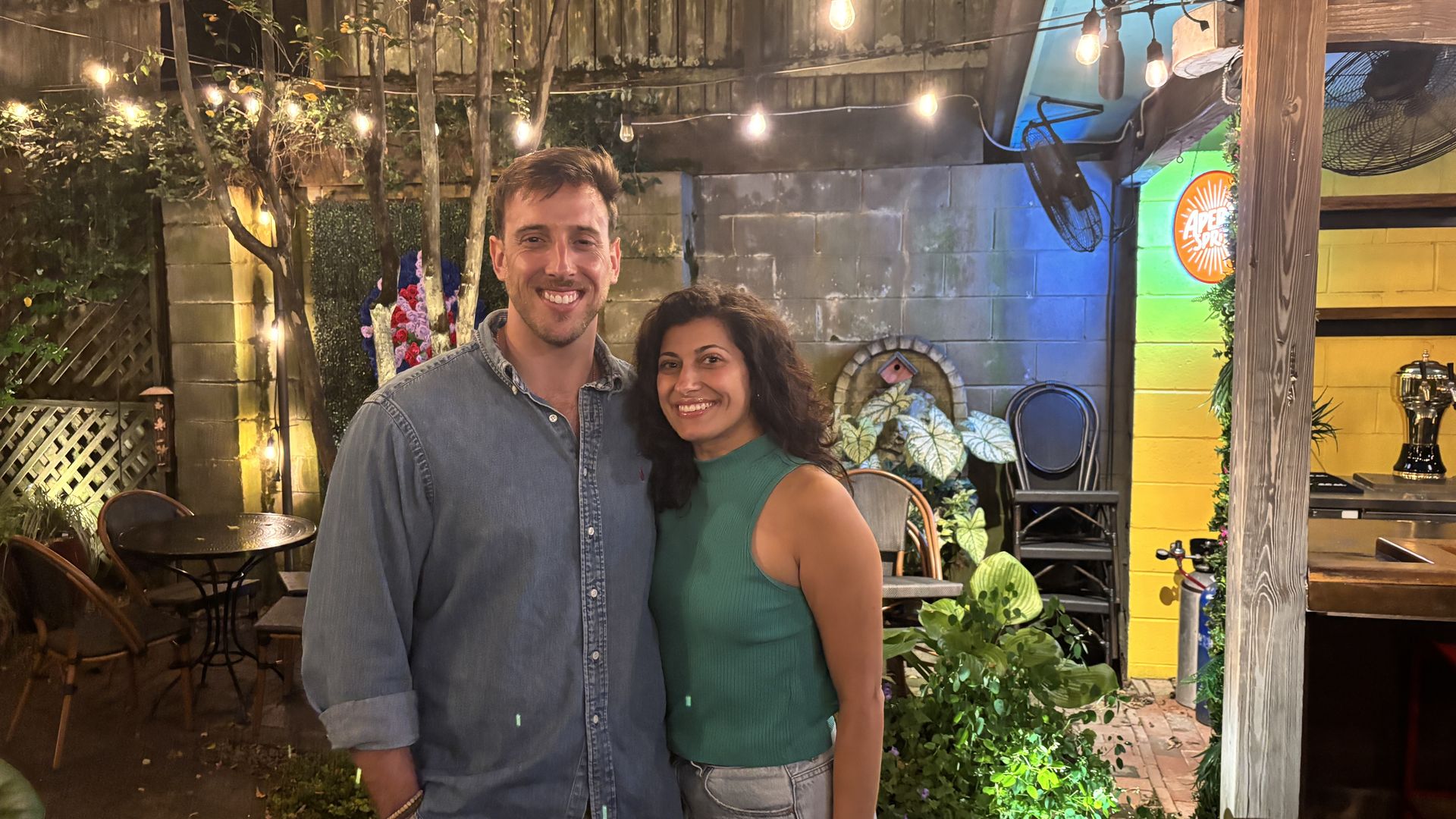 Smiling man in denim shirt and woman in green sleeveless top stand close in cozy outdoor patio with string lights, plants, and yellow and green walls at night.