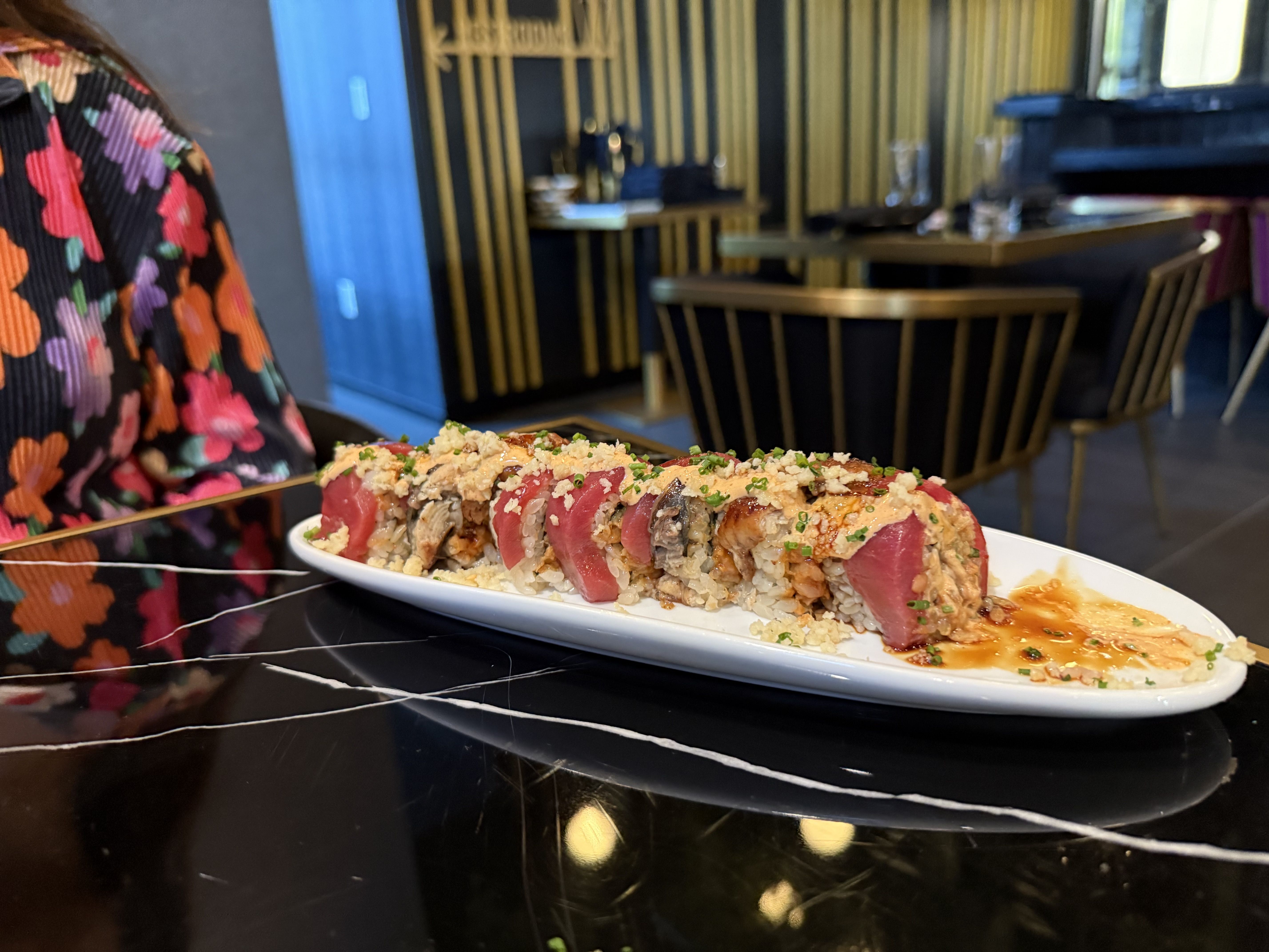 Plate of sushi rolls with pink fish, rice, sauce, and green garnish on a black marble table in a modern restaurant with gold and black chairs and floral shirt visible.